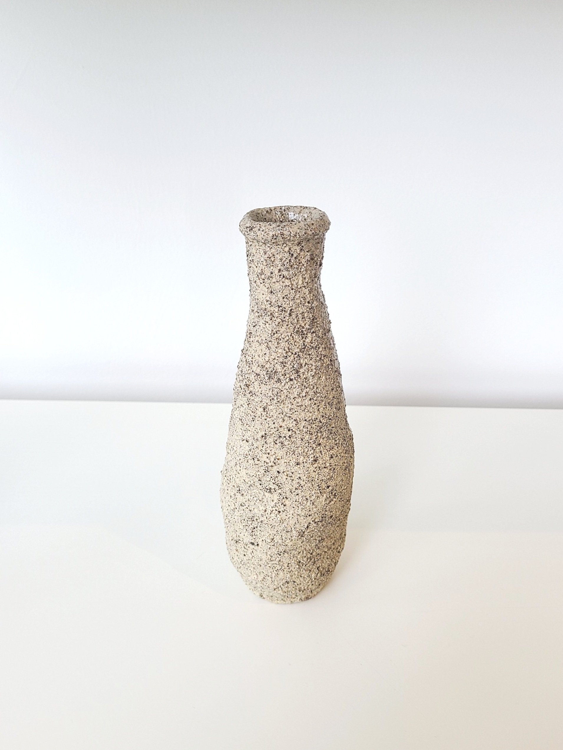 A tall, narrow, textured stone vase with a rough, speckled surface, set against a plain white background.