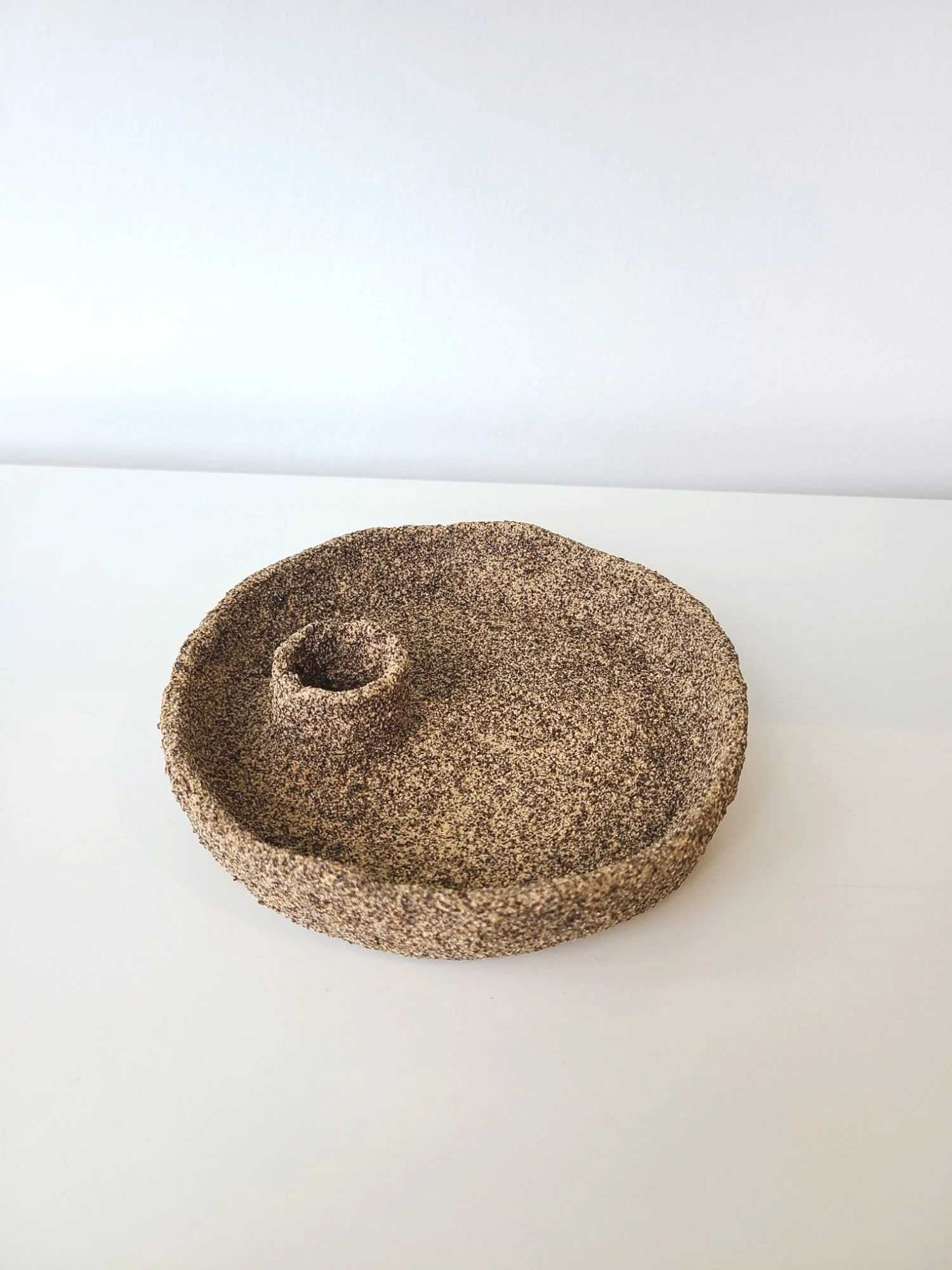 A round, textured, brown object with a small spiral indentation on one side, placed on a white surface in front of a white background.