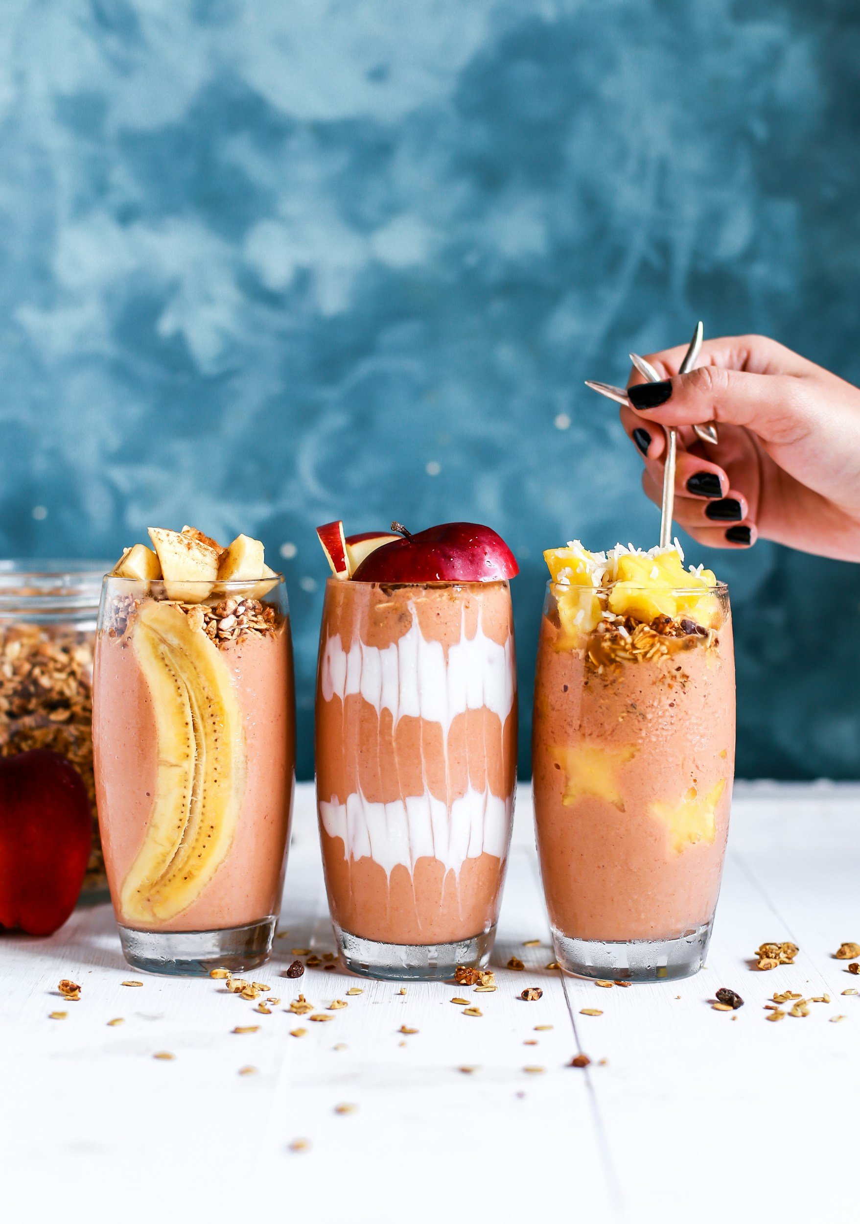 Three fruit smoothies in tall glasses topped with fruits and granola on a white surface, with a blue background.