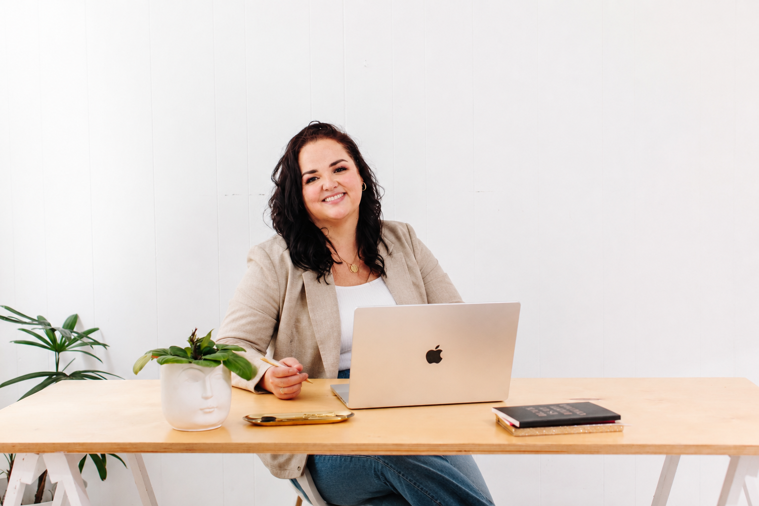 Smiling woman with dark wavy hair sitting at a desk with a laptop, a potted plant, and notebooks in a bright, minimal office.