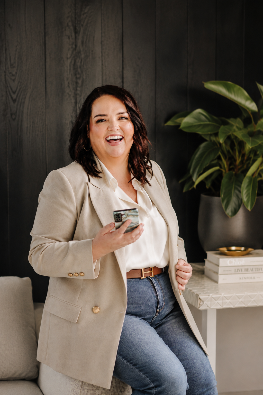 Woman with dark wavy hair, wearing a beige blazer, white blouse, and jeans, smiling and holding a smartphone while sitting on a sofa in a modern living room with a large potted plant and decorative books.