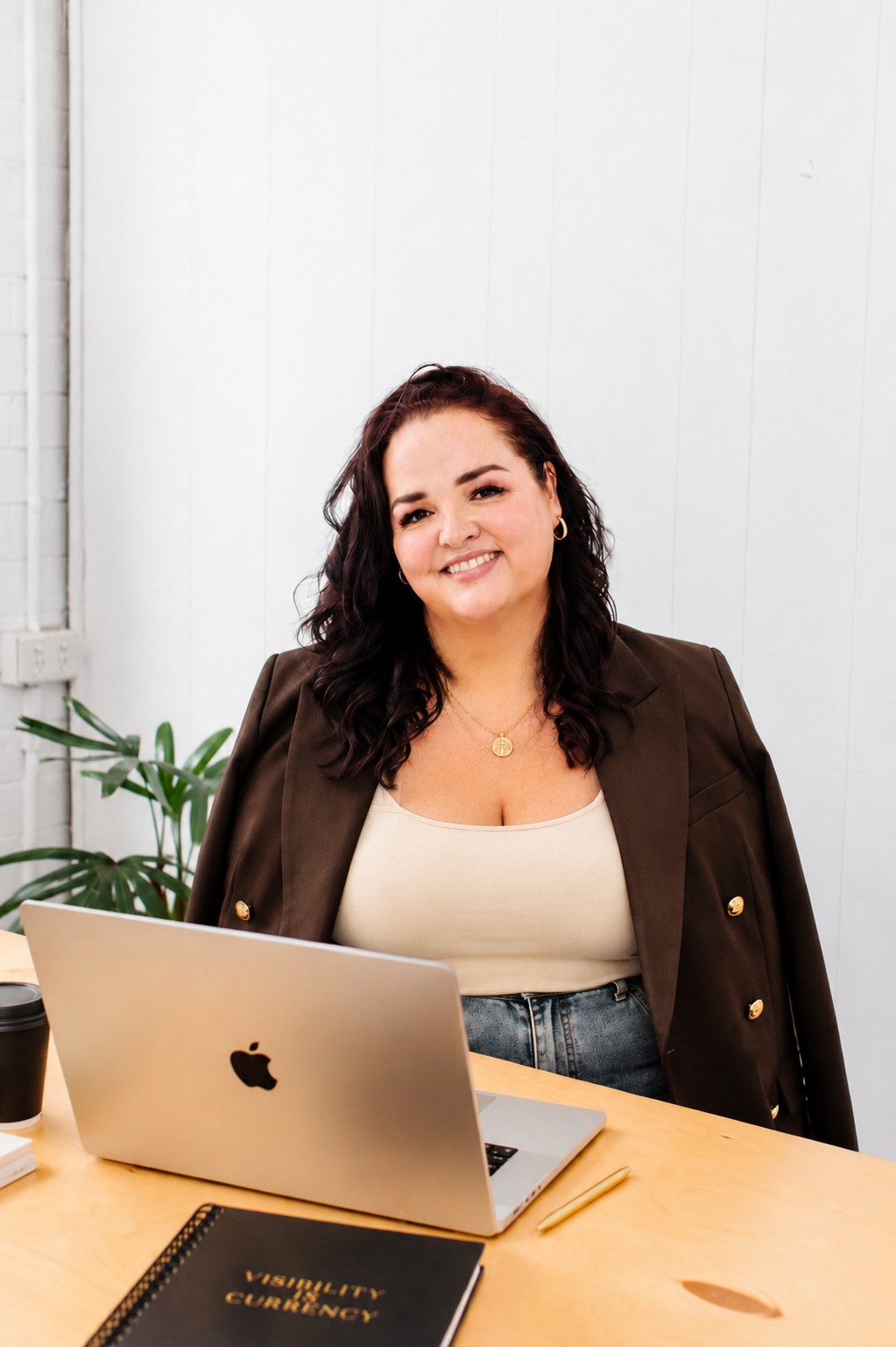 A woman with dark, wavy hair smiling at the camera, sitting at a wooden desk with a laptop, notebook, and a coffee cup, in a bright office with white paneled walls and a green plant in the background.