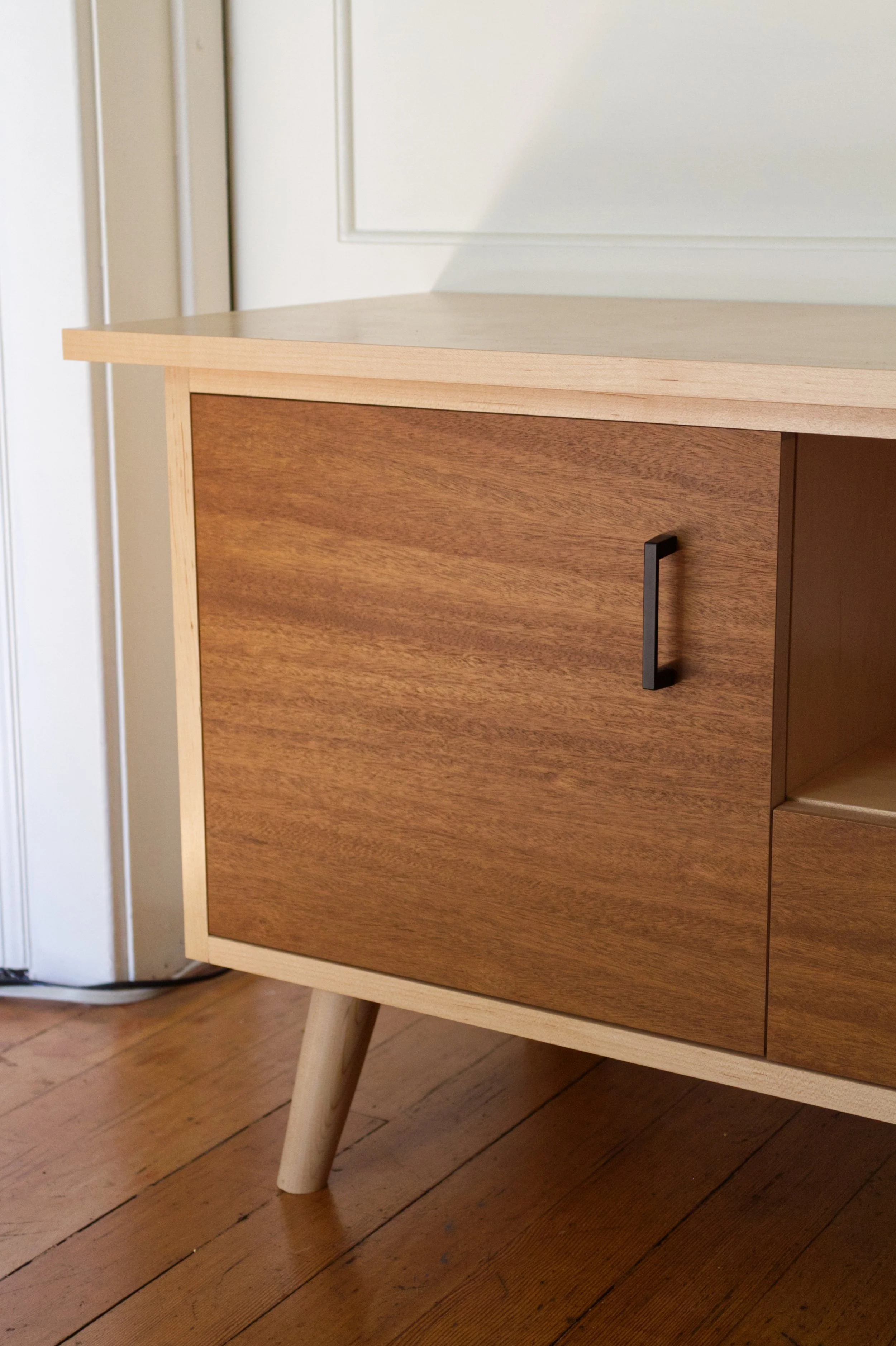 Close-up of a modern wooden cabinet with a light top and darker main body, black handle, wooden legs, and a hardwood floor beneath.