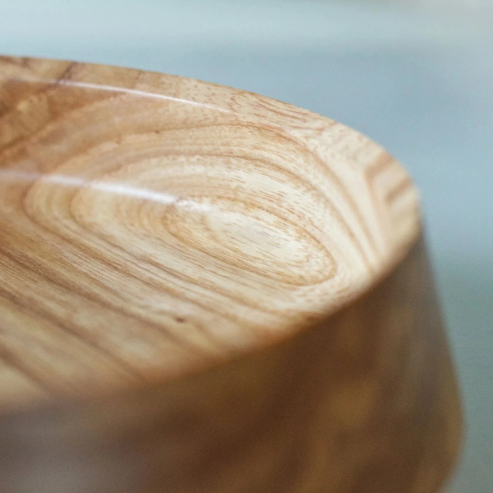 Close-up of a wooden object with a smooth, rounded edge, showing natural wood grain patterns, against a light blue background.