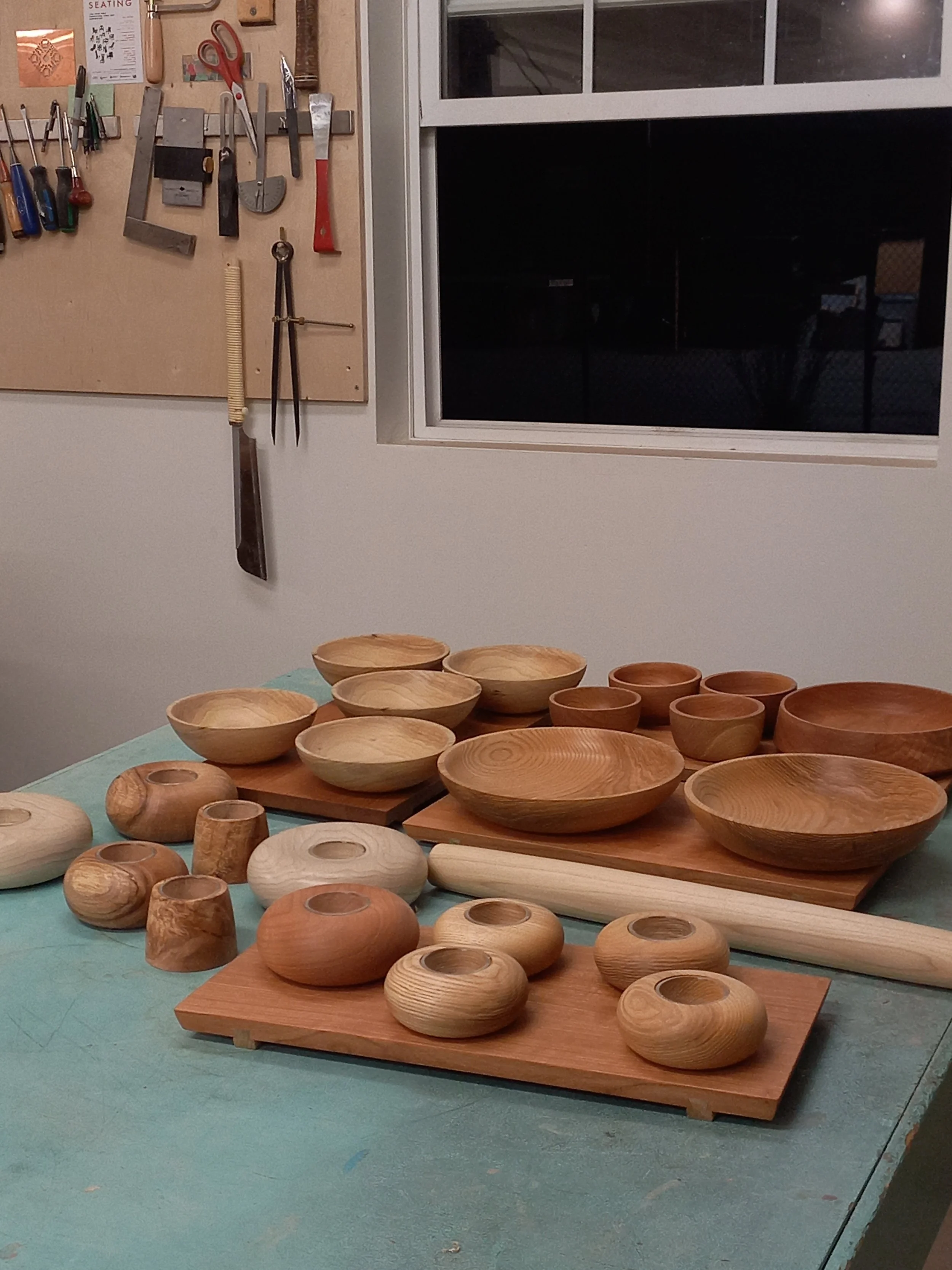Various unfinished wooden bowls and cups arranged on wooden boards and a table in a woodworking workshop. In the background, tools such as scissors, chisels, and files hang on a corkboard and wall.