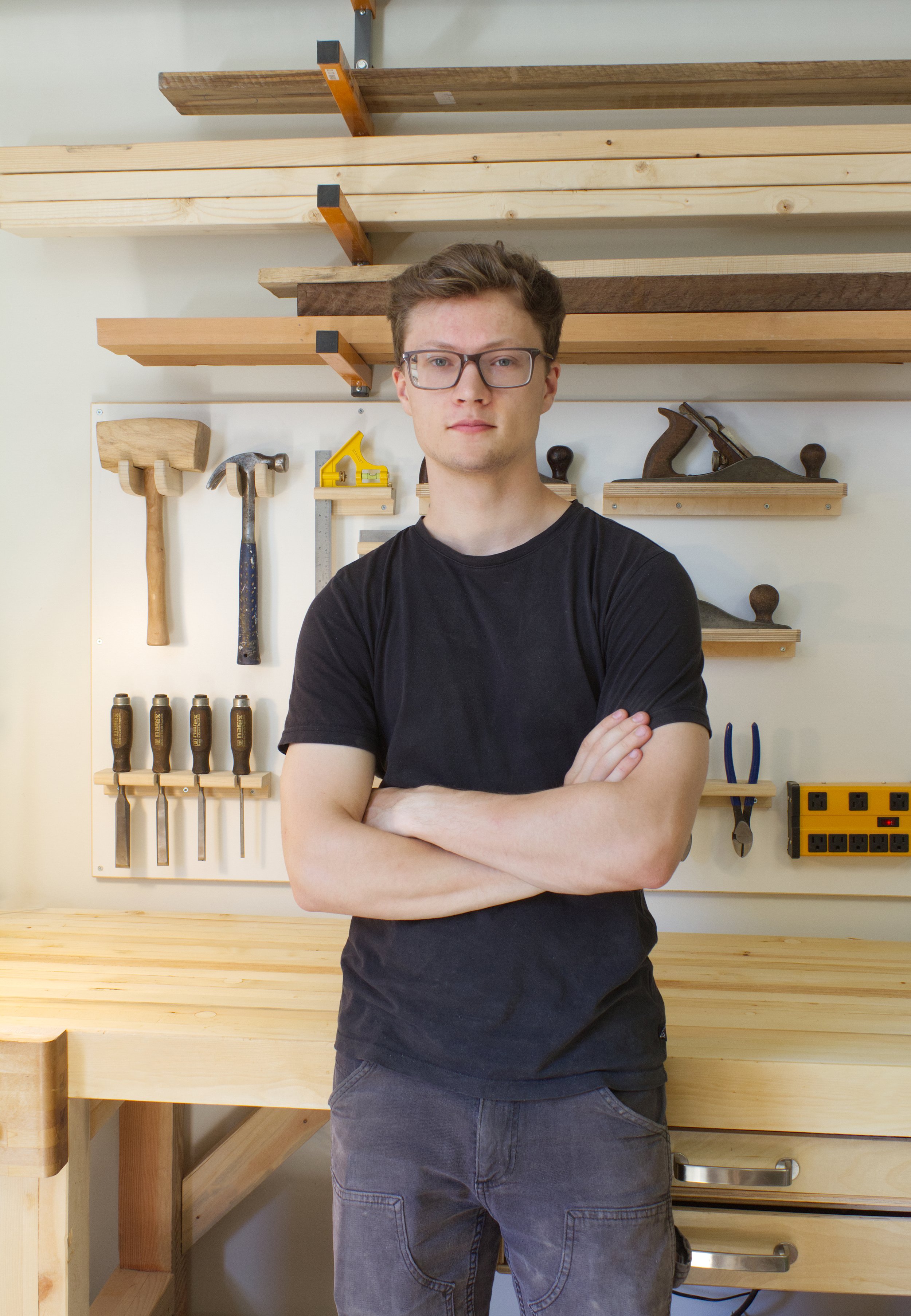 Sam Johns standing in a woodworking workshop. Behind him are hand tools such as hammers, a saw, planes, clamps, and a power strip.