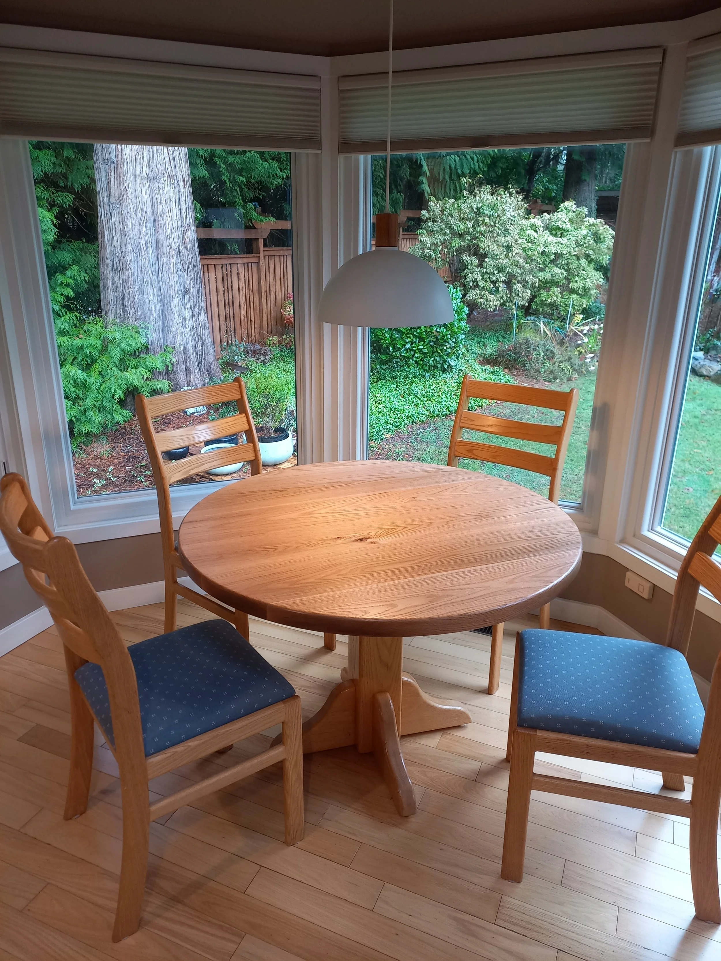 A round wooden dining table surrounded by four chairs with blue upholstered seats, situated in front of large windows overlooking a garden with trees and bushes.