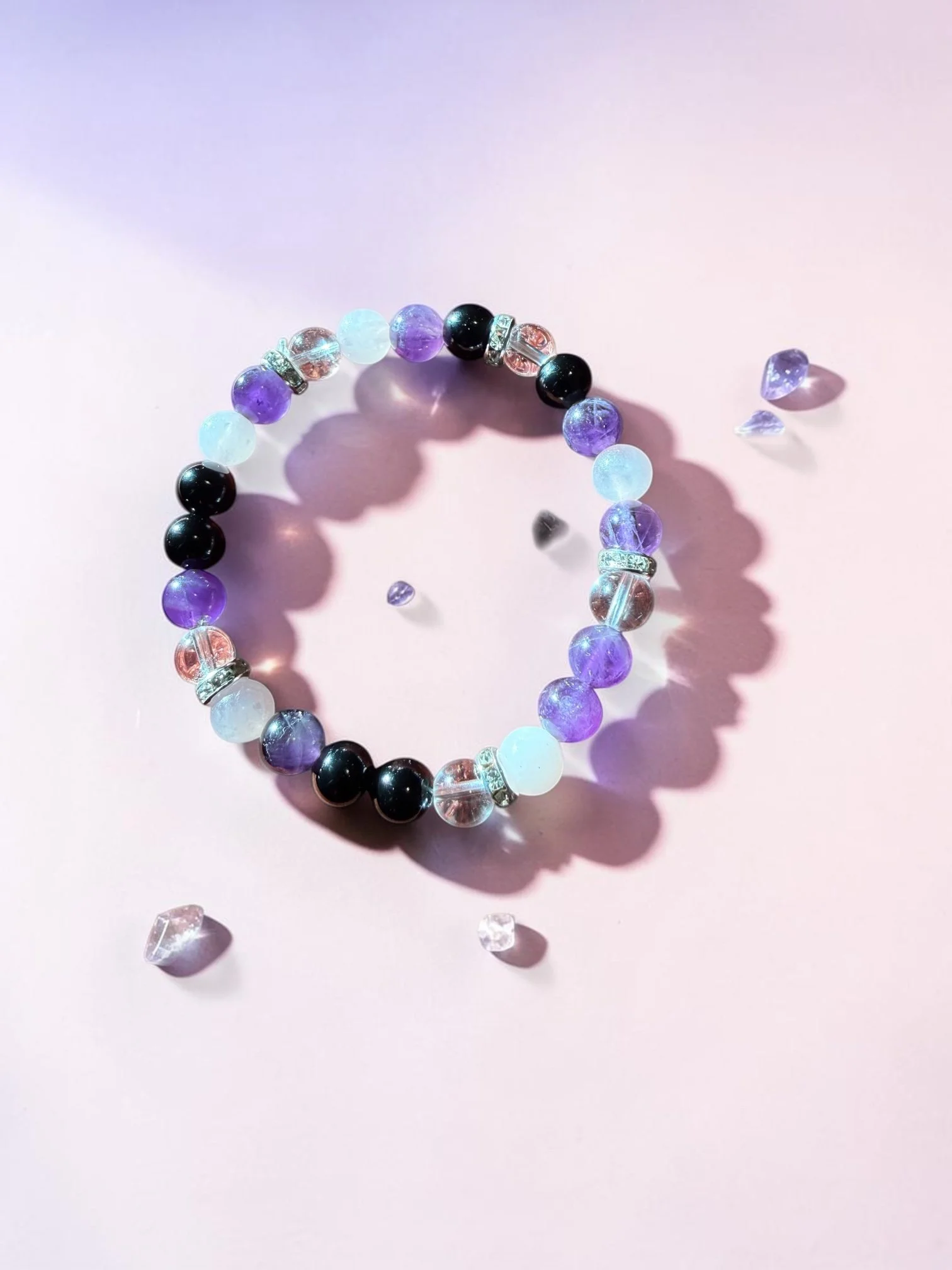 Close-up of a beaded bracelet with purple, black, white, and clear beads on a light pink surface, with scattered loose beads around it.