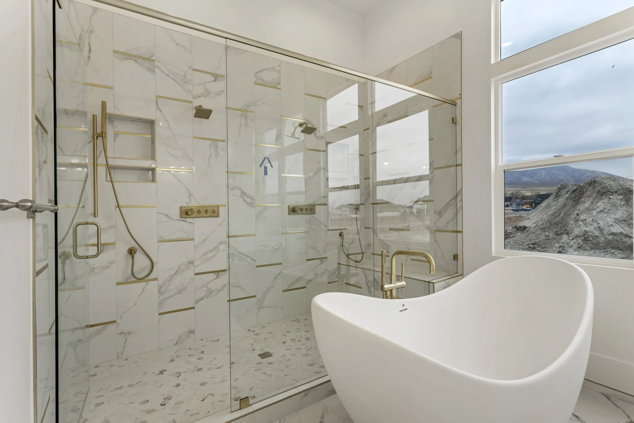 Modern bathroom with a freestanding bathtub and a glass-enclosed shower with marble and gold accents, large windows showing a mountain landscape.