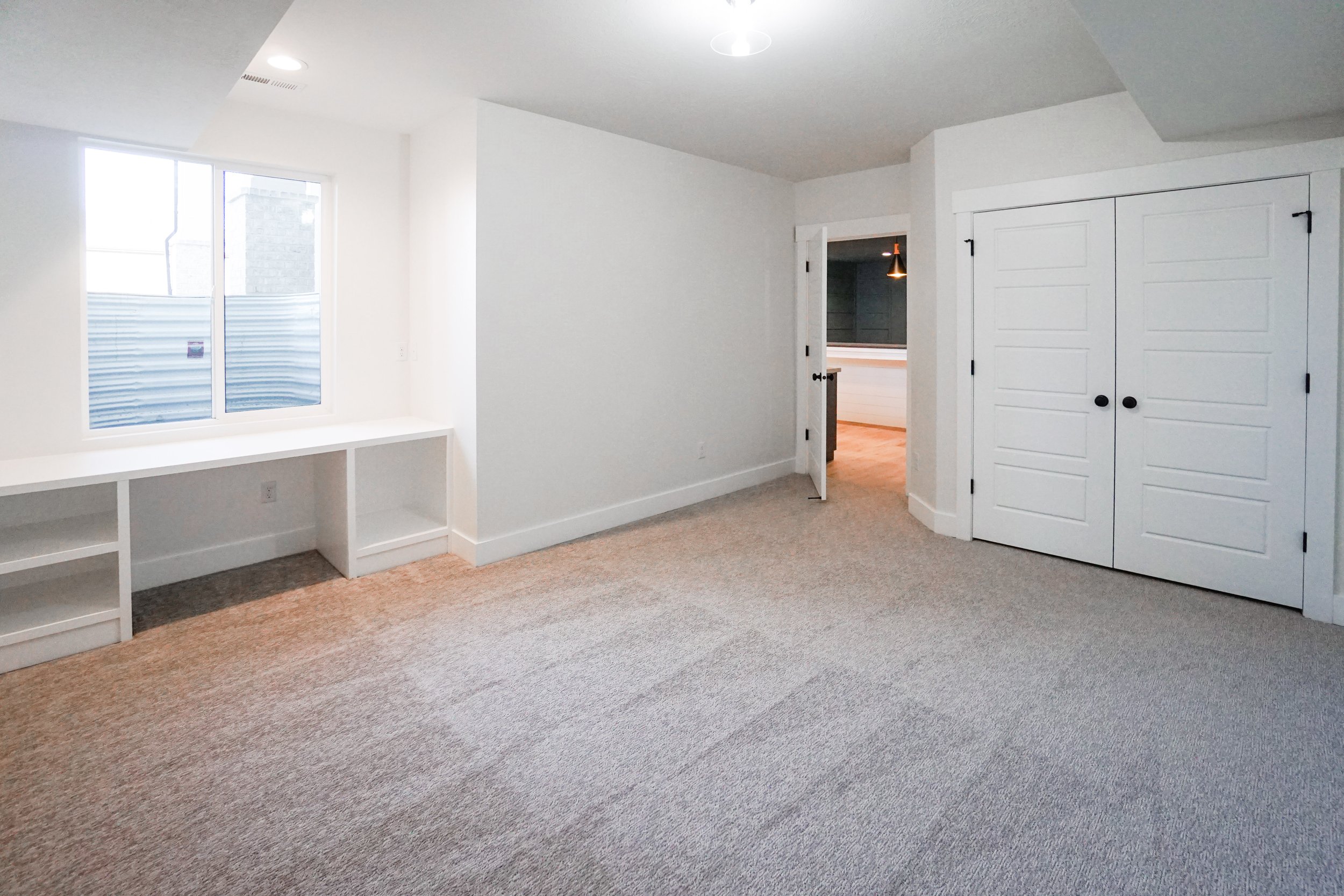 Empty room with white walls, a window, built-in white shelves, beige carpet, and a closet with double doors.