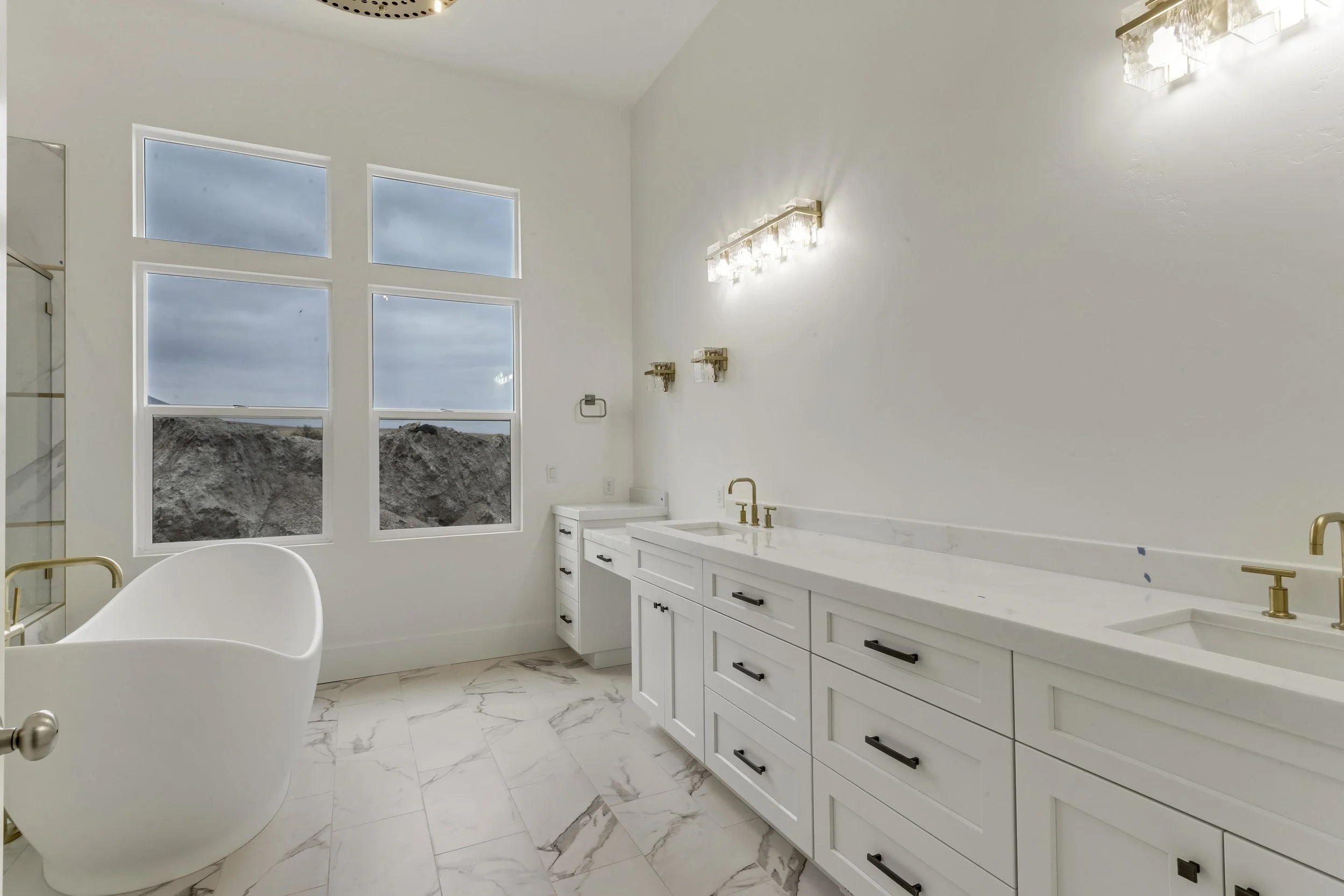 Modern bathroom with white cabinetry, gold fixtures, a floating vanity, marble flooring, and large windows showing a landscape with cloudy sky and rocky terrain.