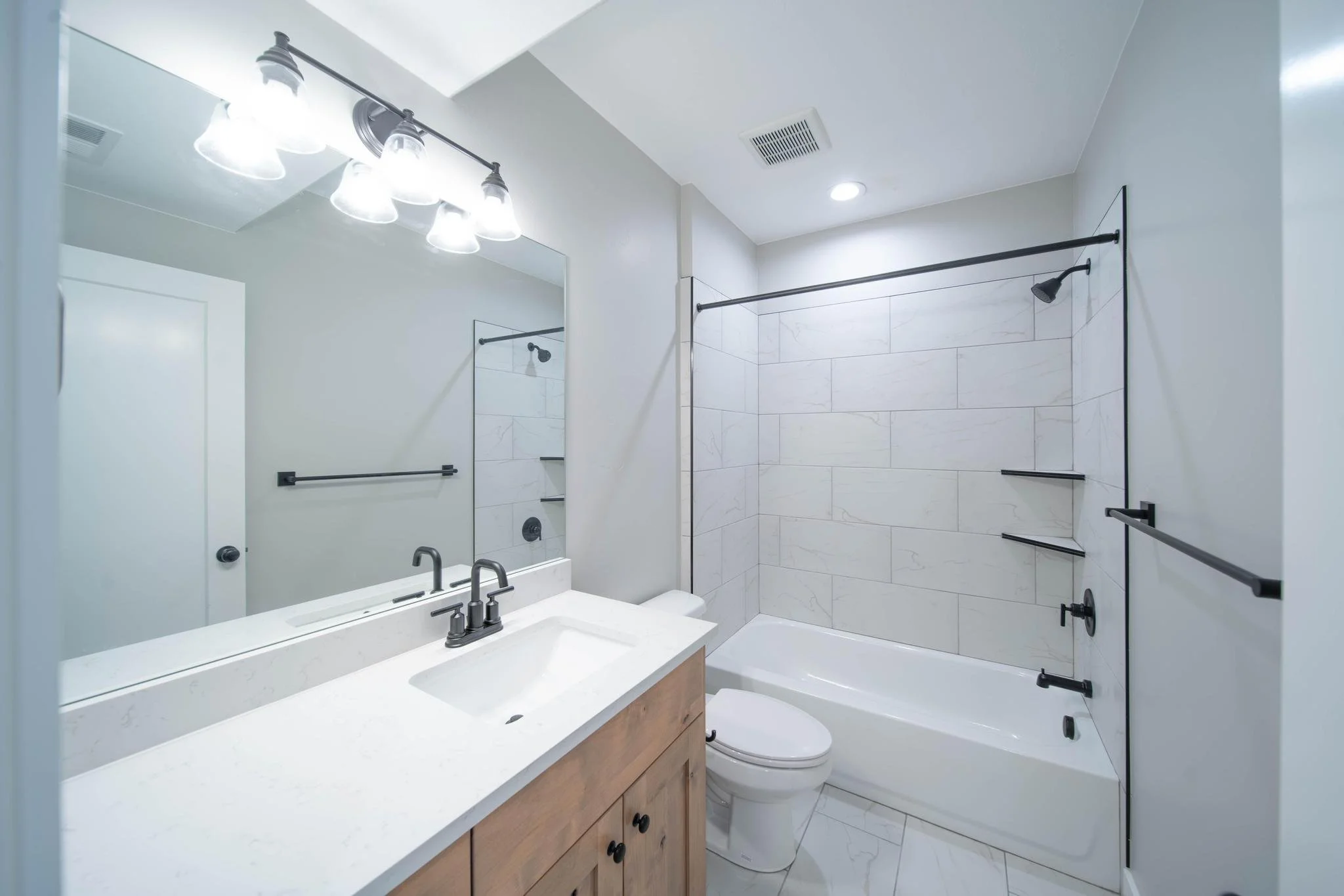 A clean, modern bathroom with a white vanity, large mirror, and a shower/tub combo with a glass door. The bathroom has white tile walls and a white floor, with black fixtures and accents.