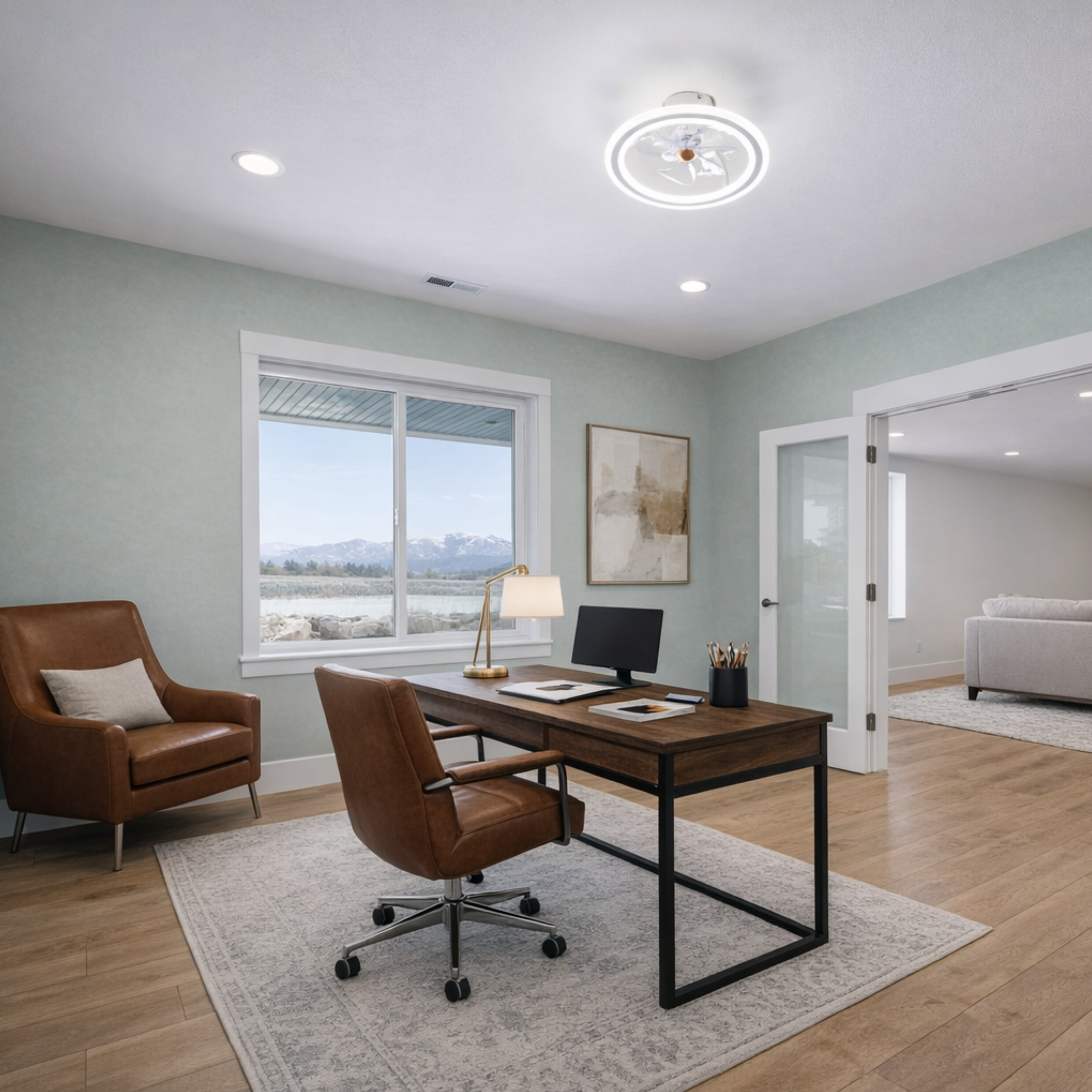 Home office with a wooden desk, black office chair, and brown leather accent chairs on a light-colored area rug. There are large windows showing a mountain landscape, a lamp, and framed artwork on the wall.