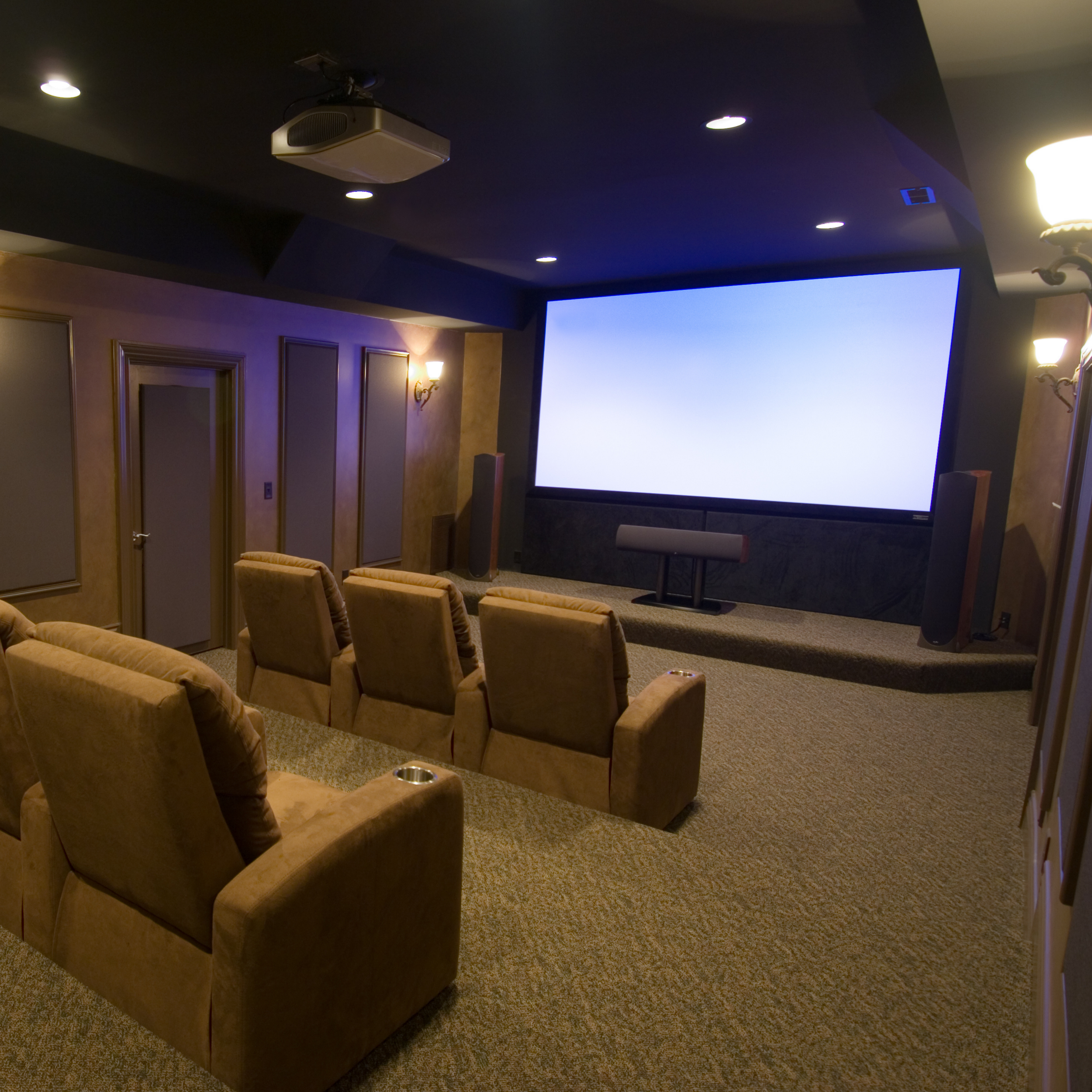 Home theater room with six beige recliner chairs facing a large blank white screen, surrounded by warm wall lighting, a ceiling-mounted projector, and a soundbar below the screen.