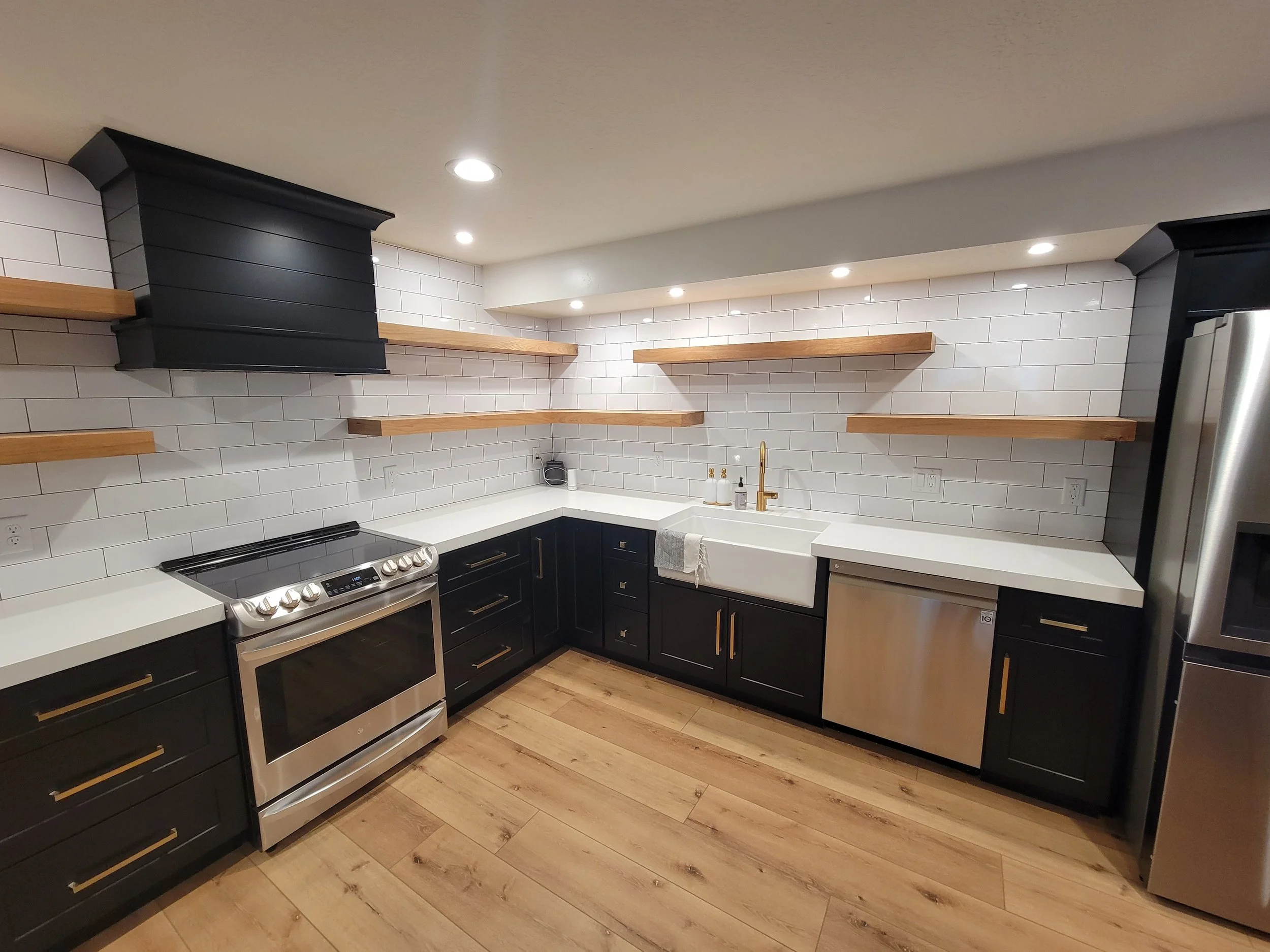 Modern kitchen with black cabinets, wooden open shelves, white subway tile backsplash, stainless steel appliances, farmhouse sink, and wood flooring.