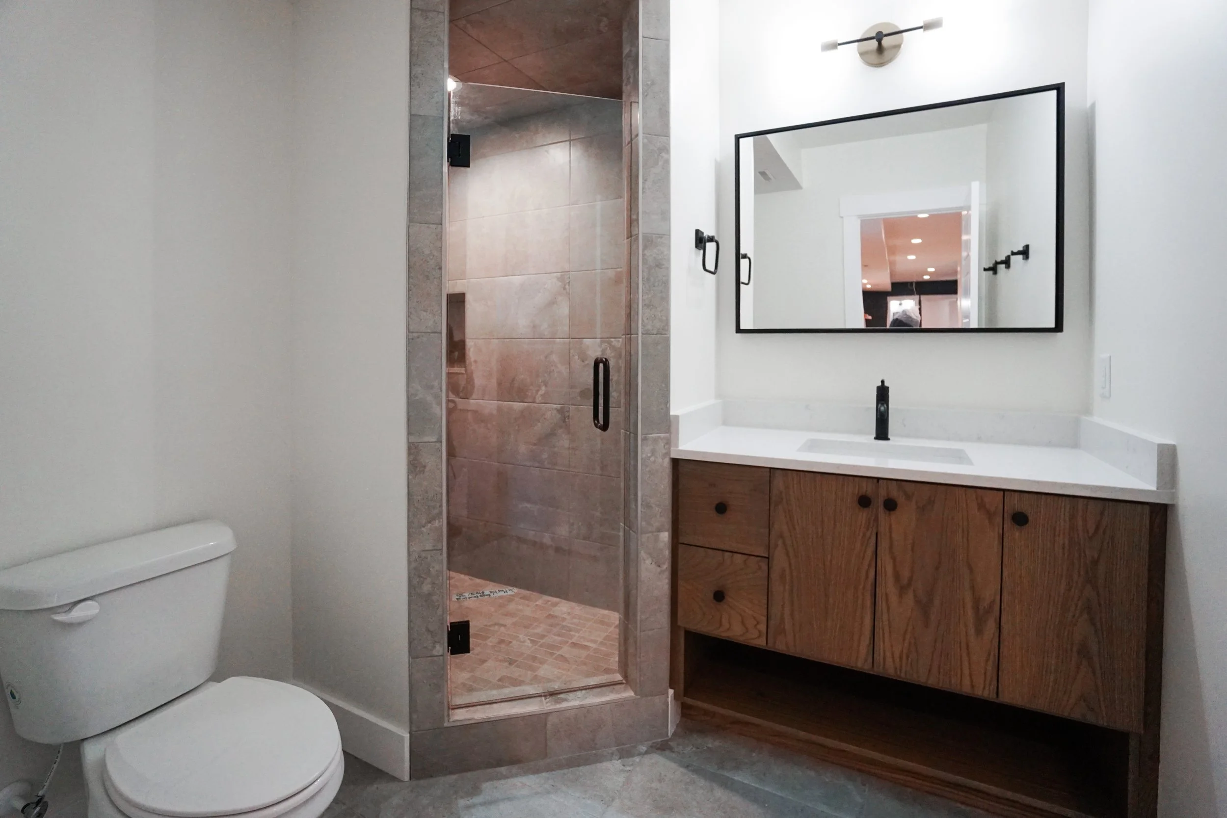 A modern bathroom featuring a white toilet, a glass shower with tan tiles, a wooden vanity with a white countertop and a black faucet, a large rectangular mirror, and minimalist black fixtures.