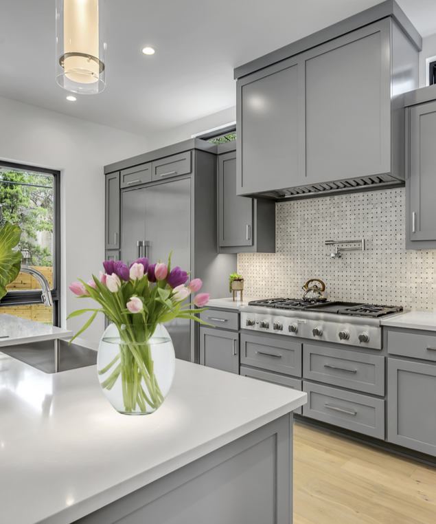 Modern kitchen with gray cabinets, white countertops, a bouquet of pink and purple tulips in a glass vase, and a gas stove with a backsplash, with a sliding glass door and greenery outside.