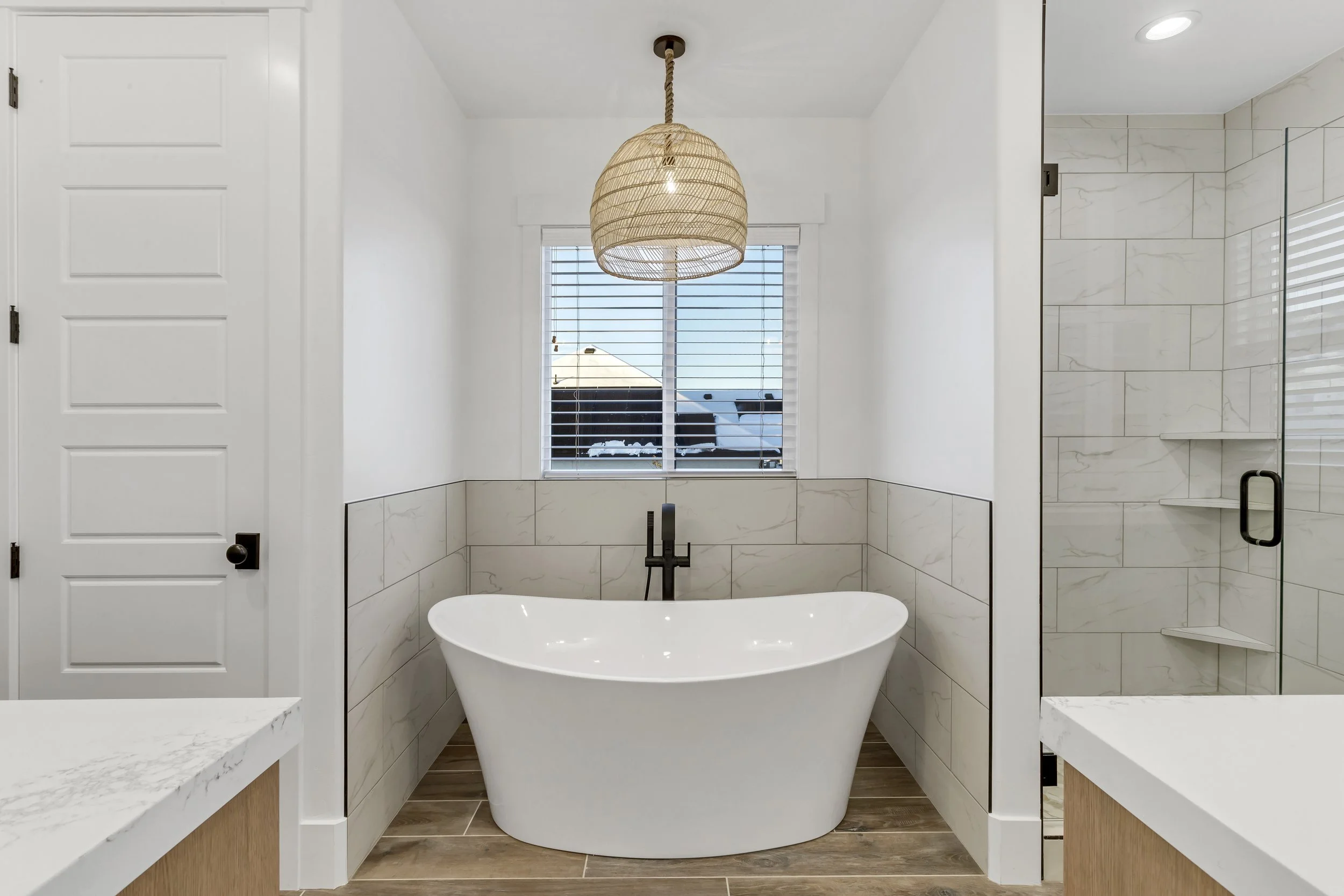 Modern bathroom with freestanding bathtub, tiled walls, glass shower enclosure, window with blinds, wooden flooring, and a woven pendant light
