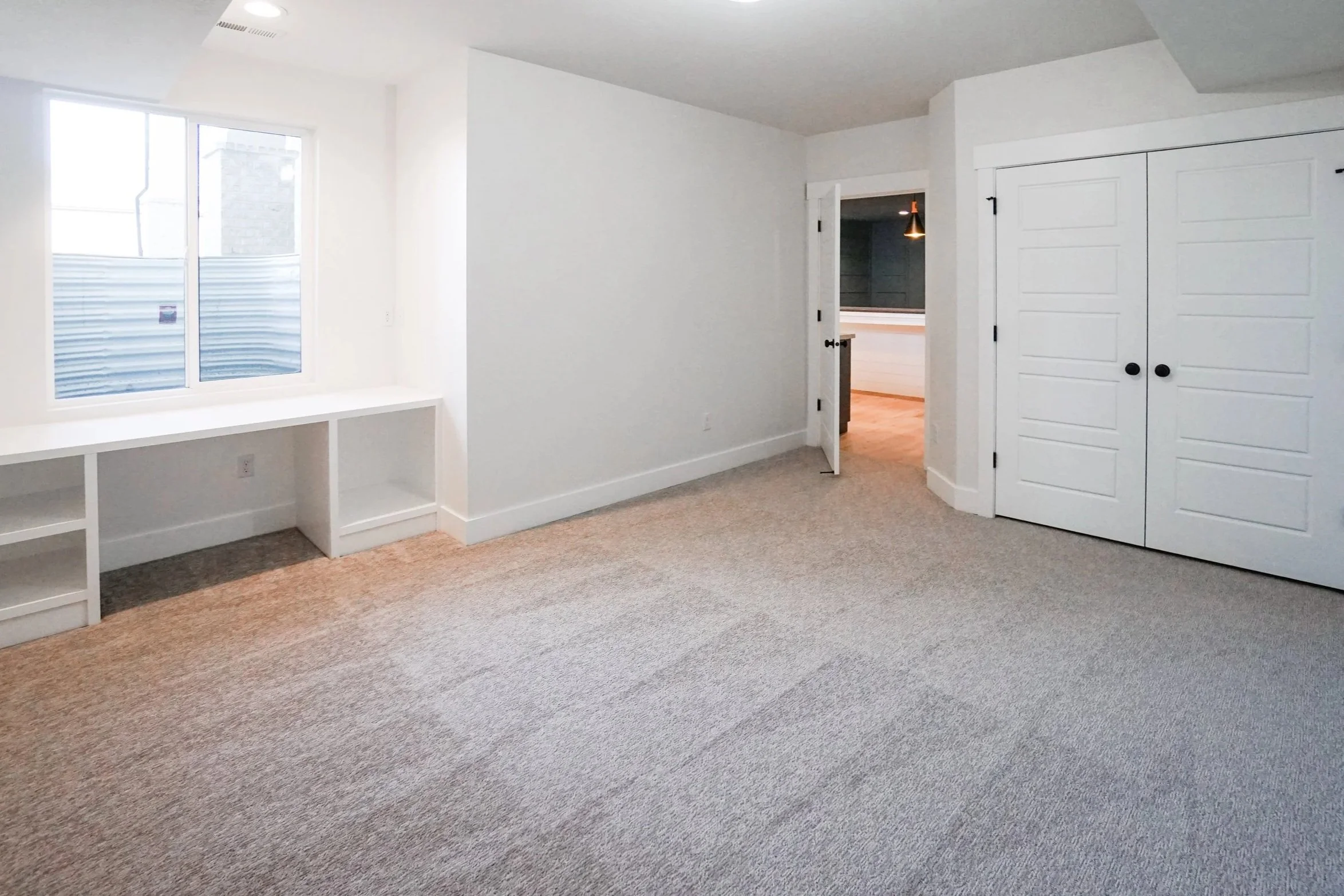 Empty room with beige carpet, white walls, a window with built-in shelves below it, a closet with white sliding doors, and a doorway leading to another room with wood flooring and hanging lights.