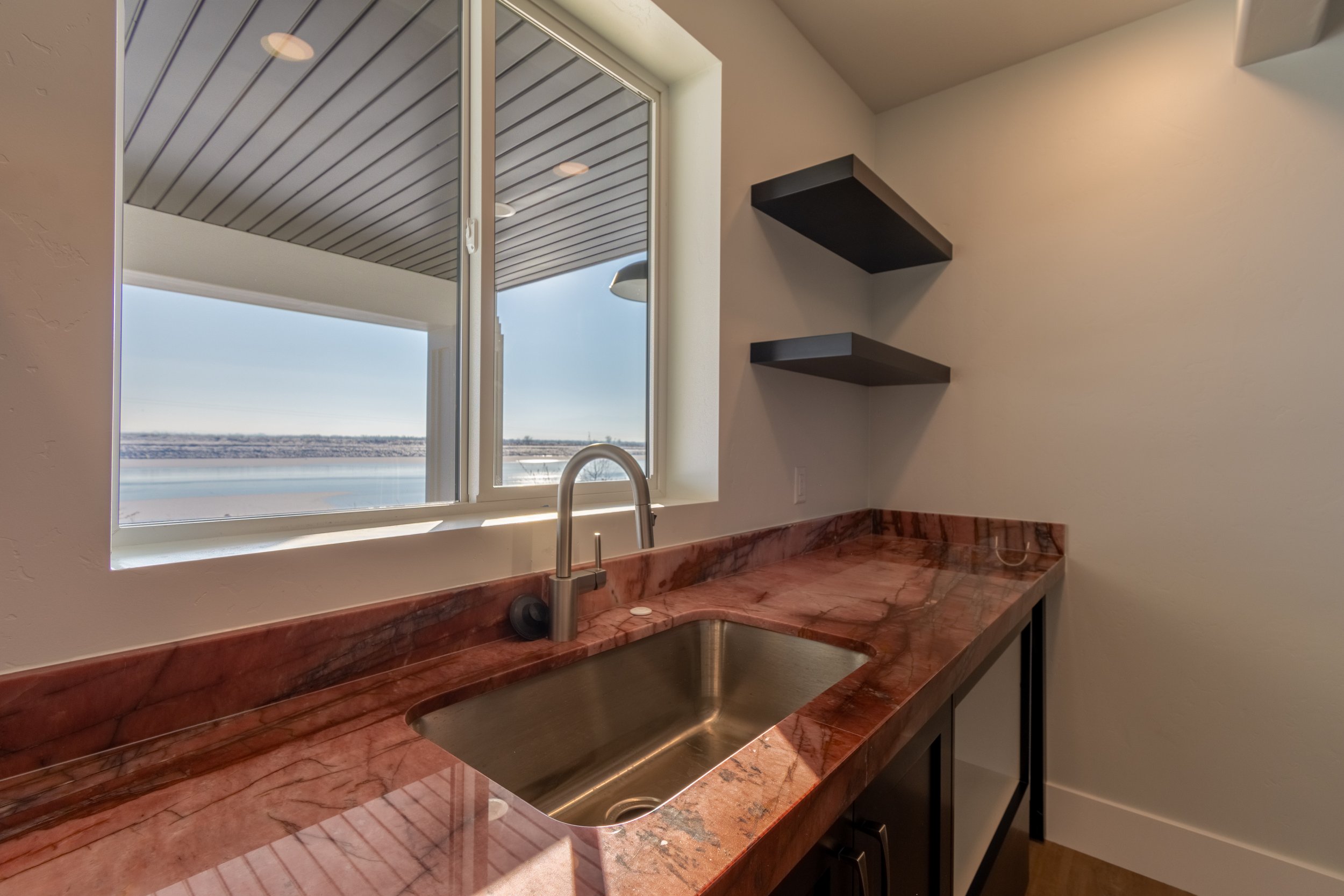 Kitchen with a red marble countertop and a stainless steel sink, overlooking a body of water through a window with sliding glass.