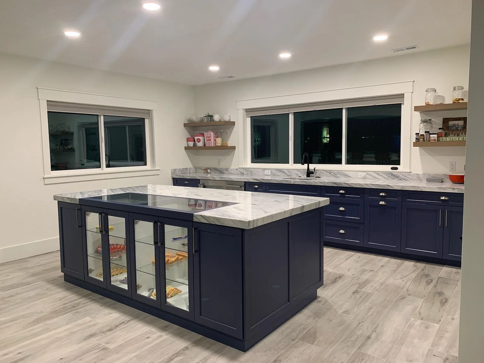 Media room snack bar kitchen with navy blue cabinets, a marble countertop island with glass display cabinets, light wood flooring, and large windows at night.