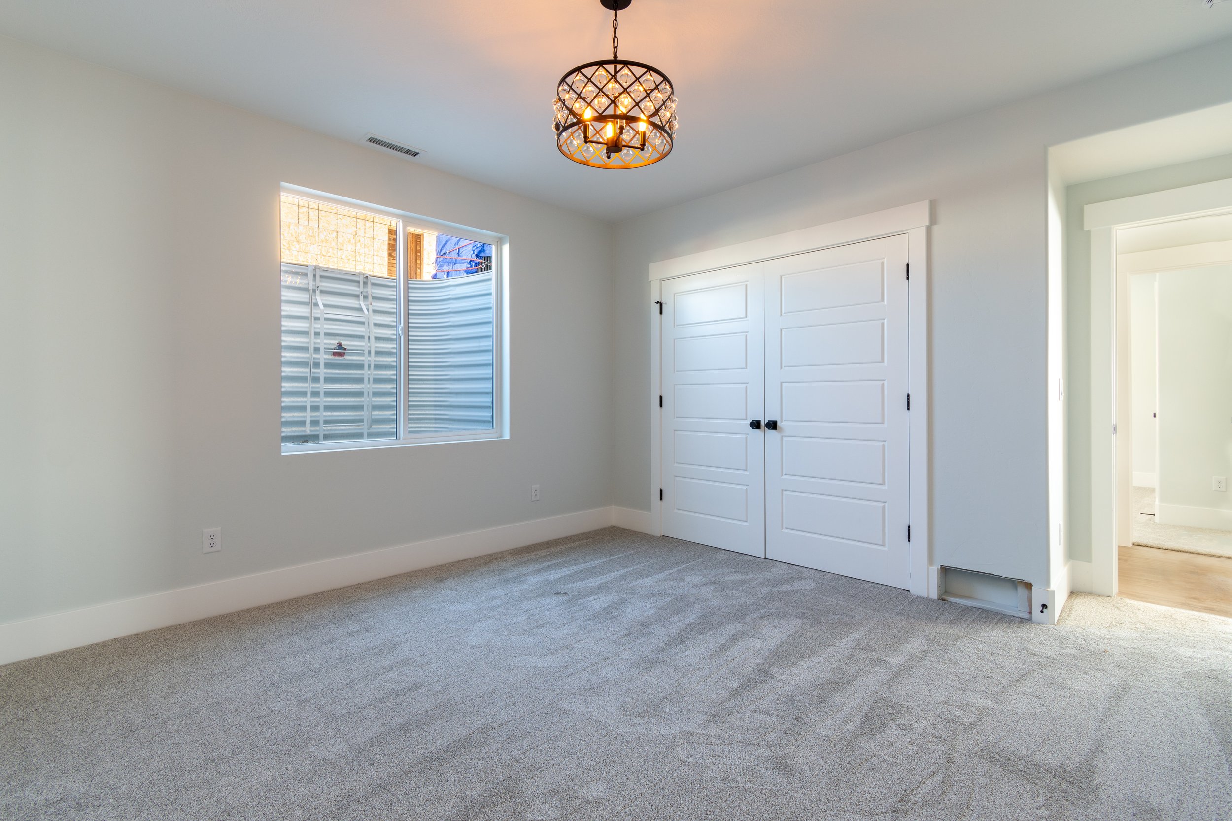 Empty room with light-colored walls, beige carpet, a window, a modern chandelier, and white double closet doors.