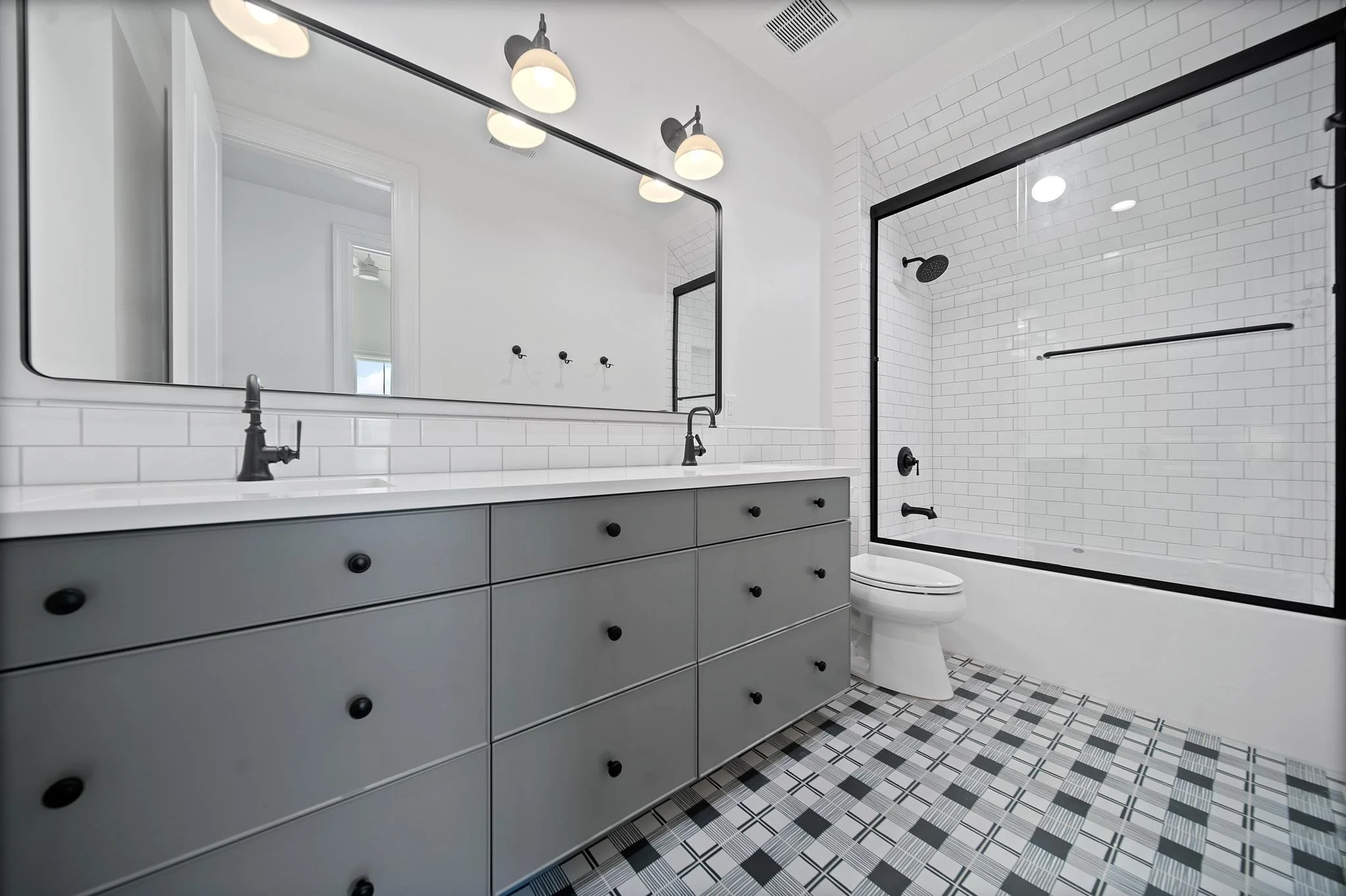 Modern bathroom with a large mirror above a gray vanity with black knobs, a white countertop, and black fixtures, a walk-in shower with a black frame, white subway tile walls, a black showerhead, and a black grab bar, and a patterned tile floor.
