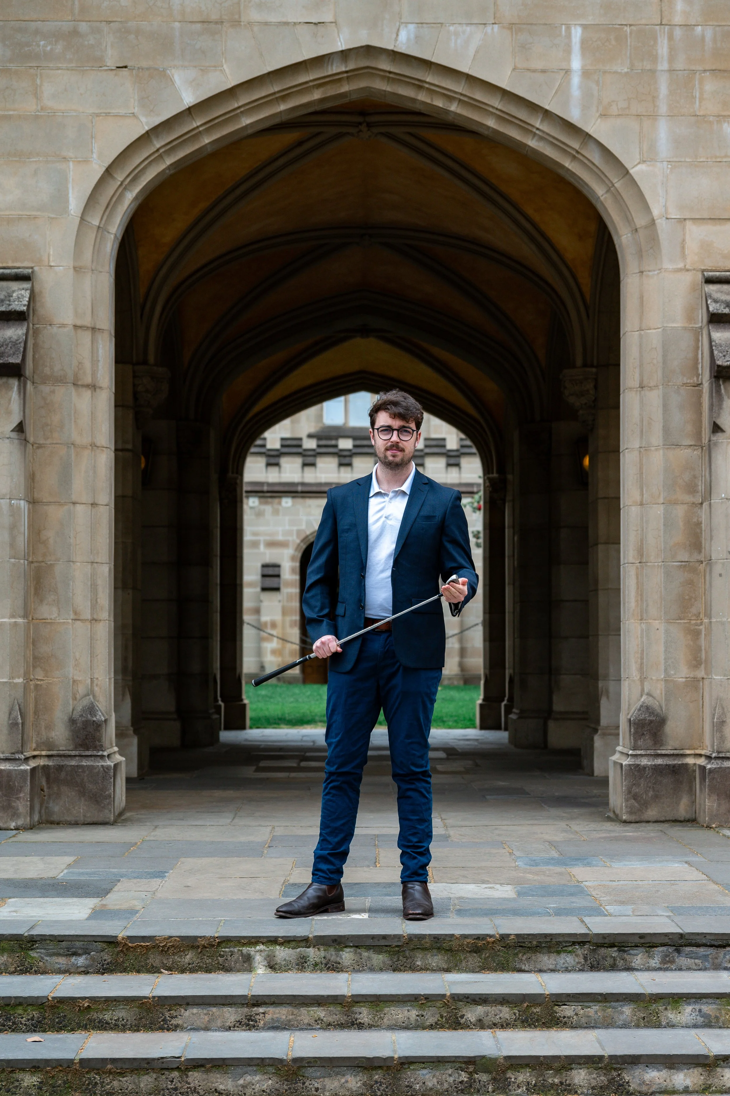 A man in a business suit holding a cane standing on stone steps in front of a historic stone building with arched entryways.