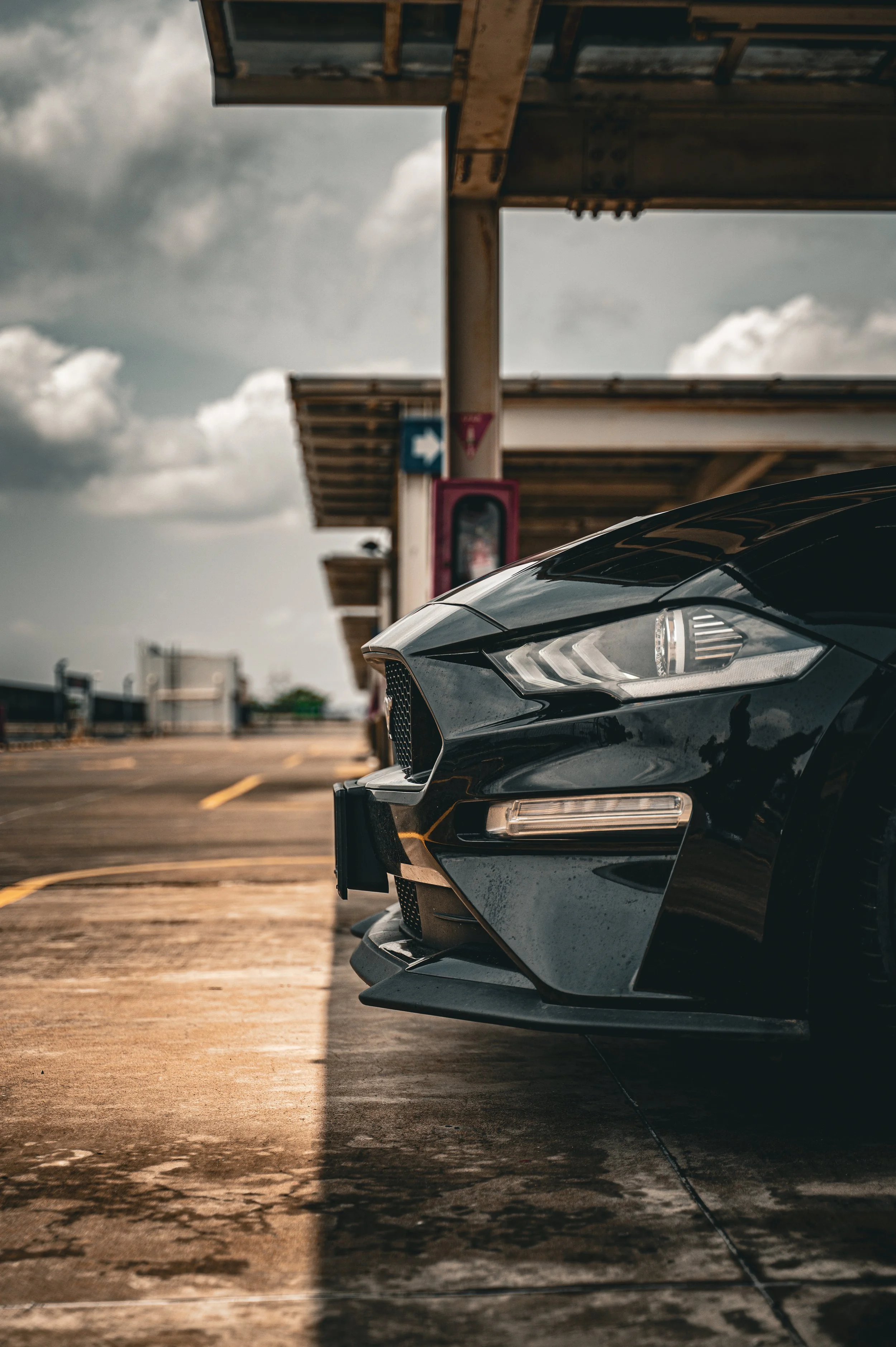 Close-up of the front end of a black sports car parked on a rooftop parking lot under a cloudy sky.