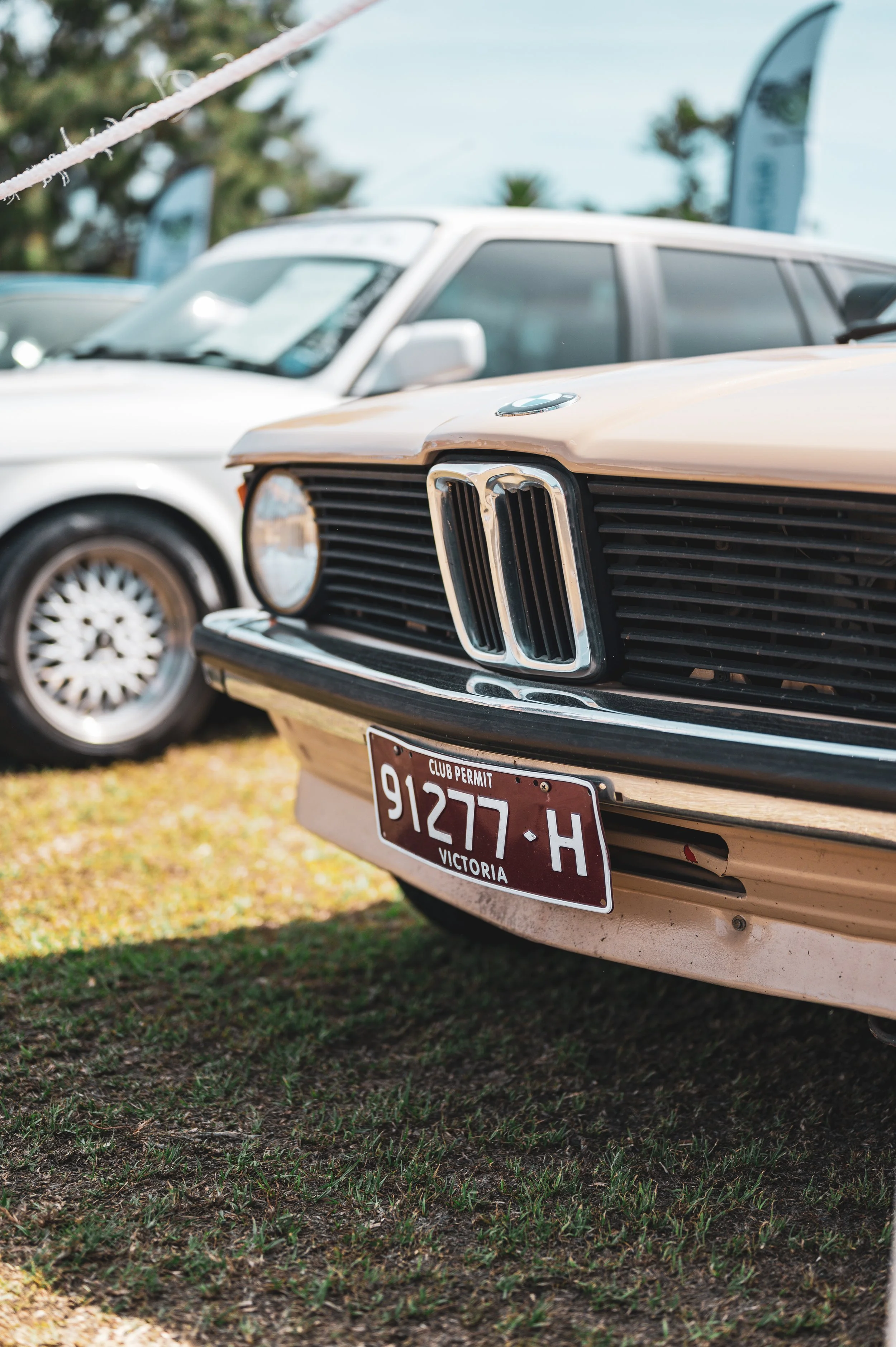 Close-up of a vintage beige BMW car with a license plate from Victoria, Australia, parked on grass during daytime, with other classic cars visible in the background.