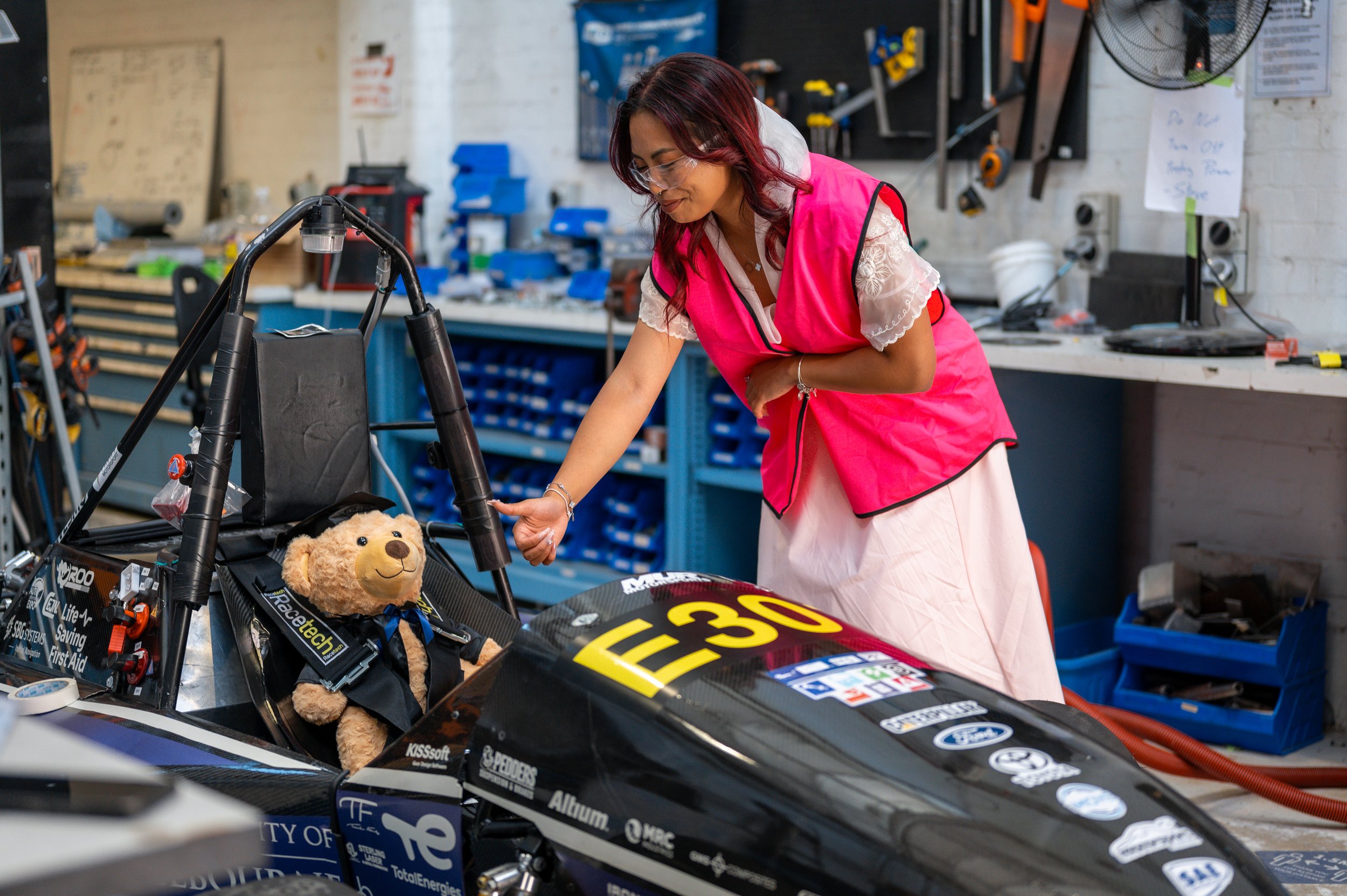 A woman in a pink vest and white dress working on a race car in a workshop, with a stuffed bear seated in the car's cockpit.