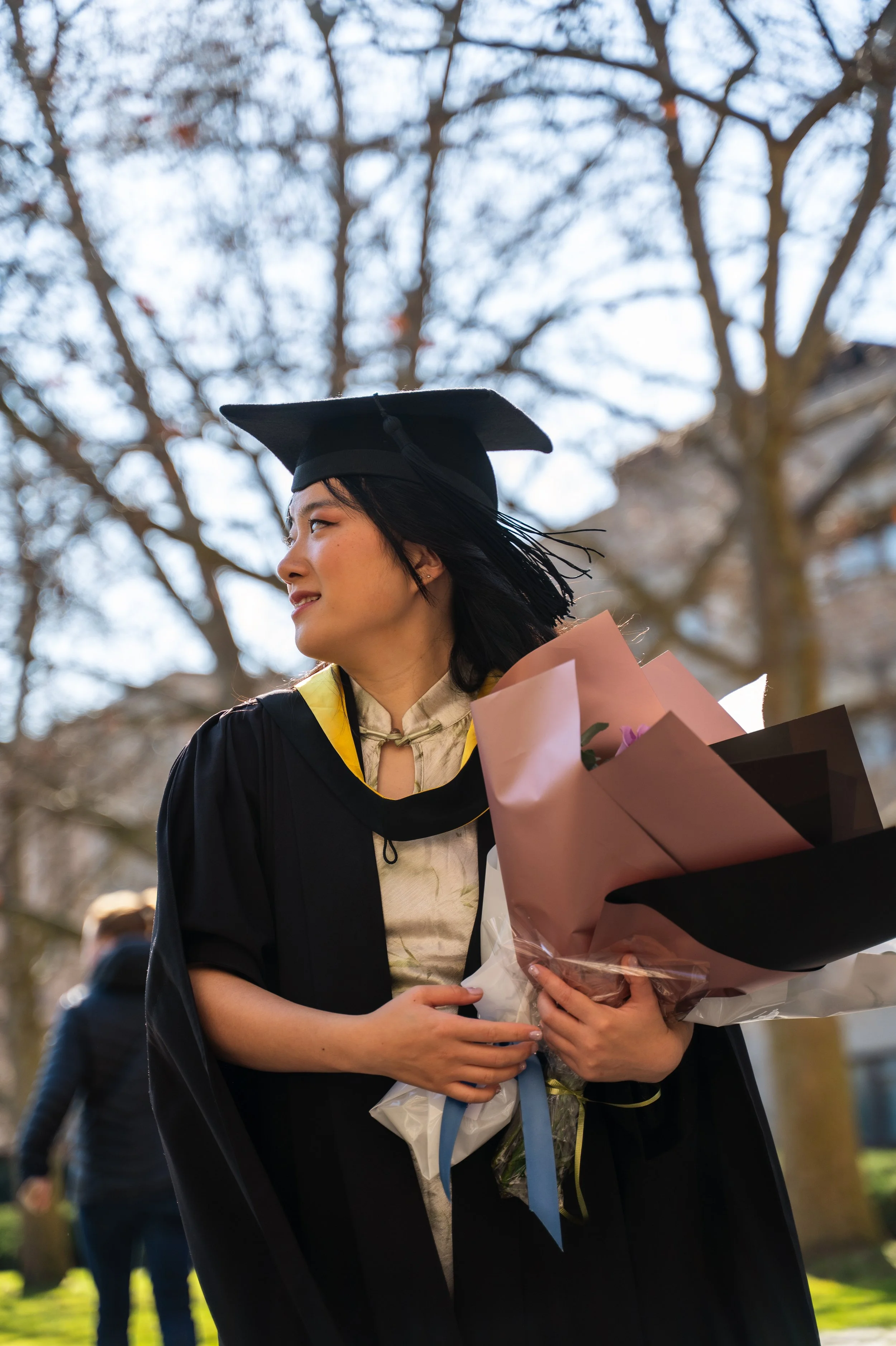 Young woman in a graduation cap and gown holding a bouquet of flowers, standing outdoors under leafless trees.