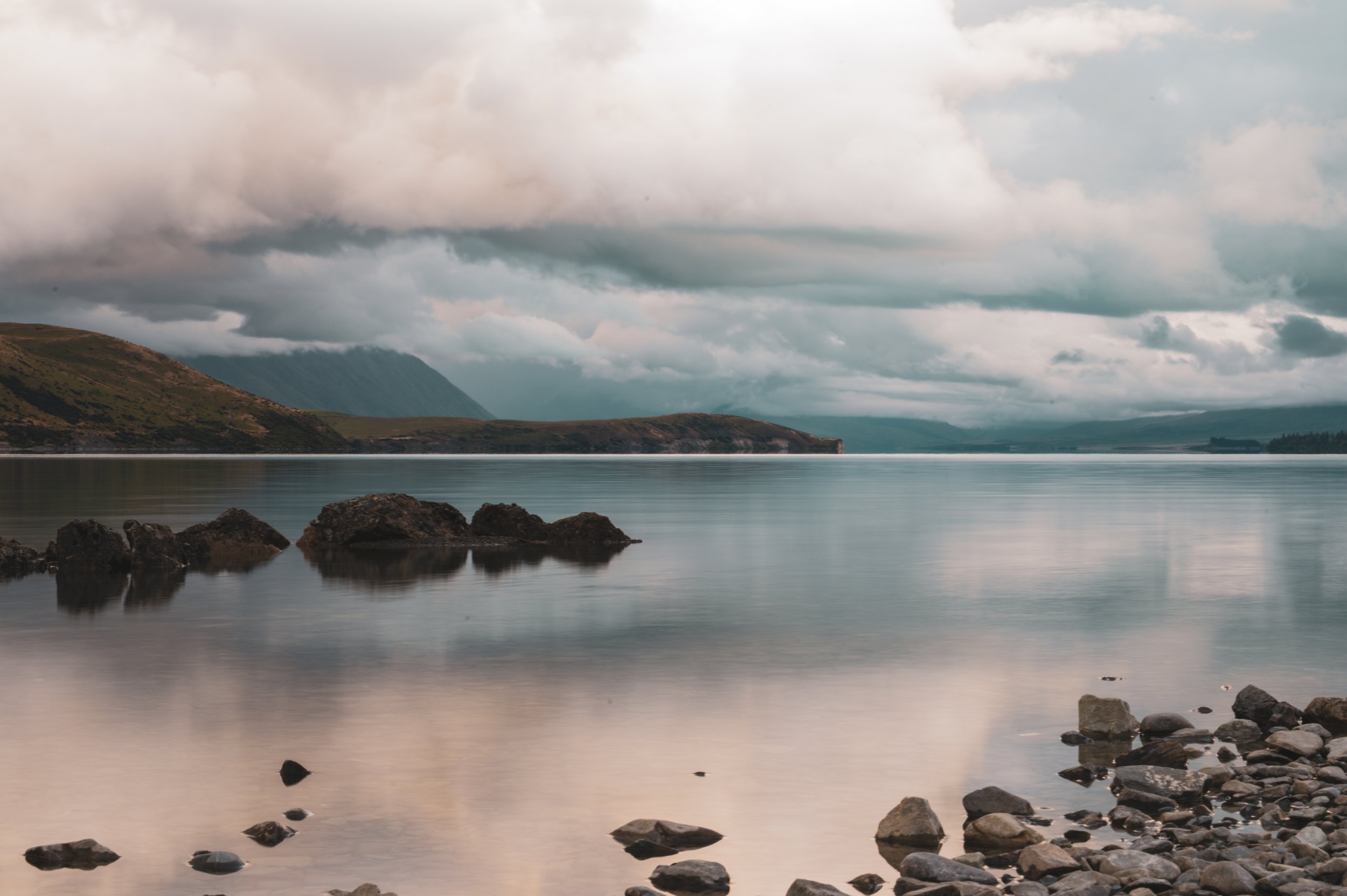 Calm lake with rocks in the foreground, distant green hills, and a cloudy sky reflected on the water.