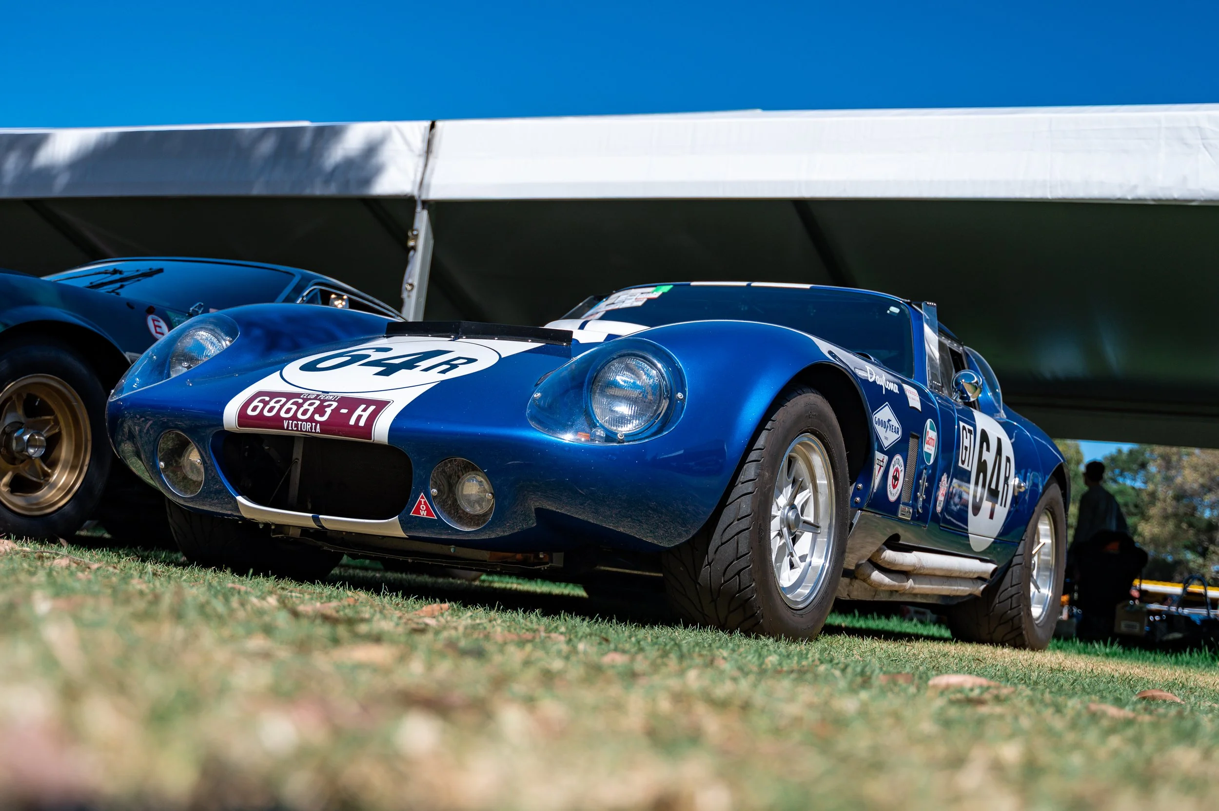 A vintage blue racing car with the number 64R on the side, parked on grass under a white canopy at a car show.