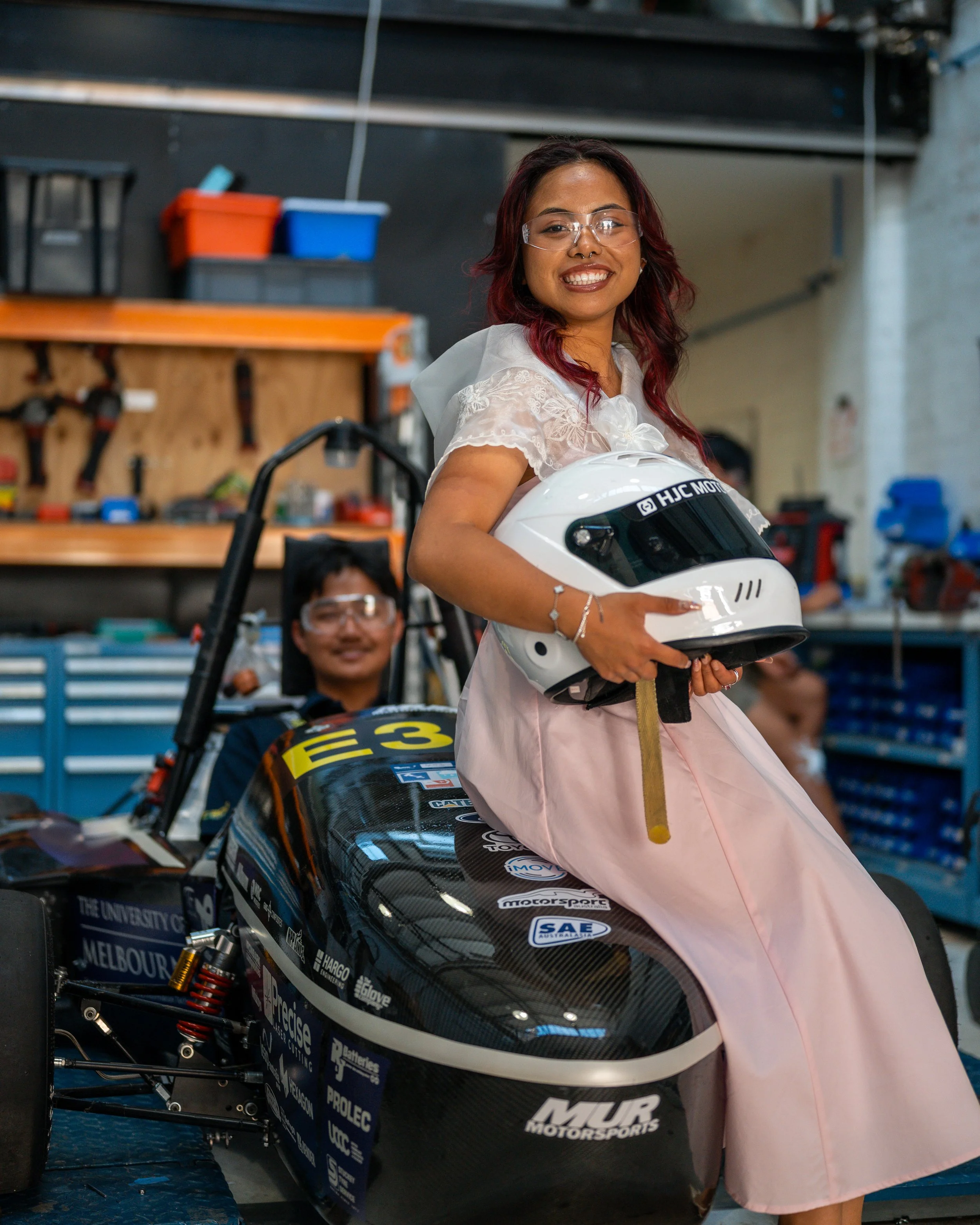 Woman holding a white motorcycle helmet sitting on a race car in a garage, with a man in the background.