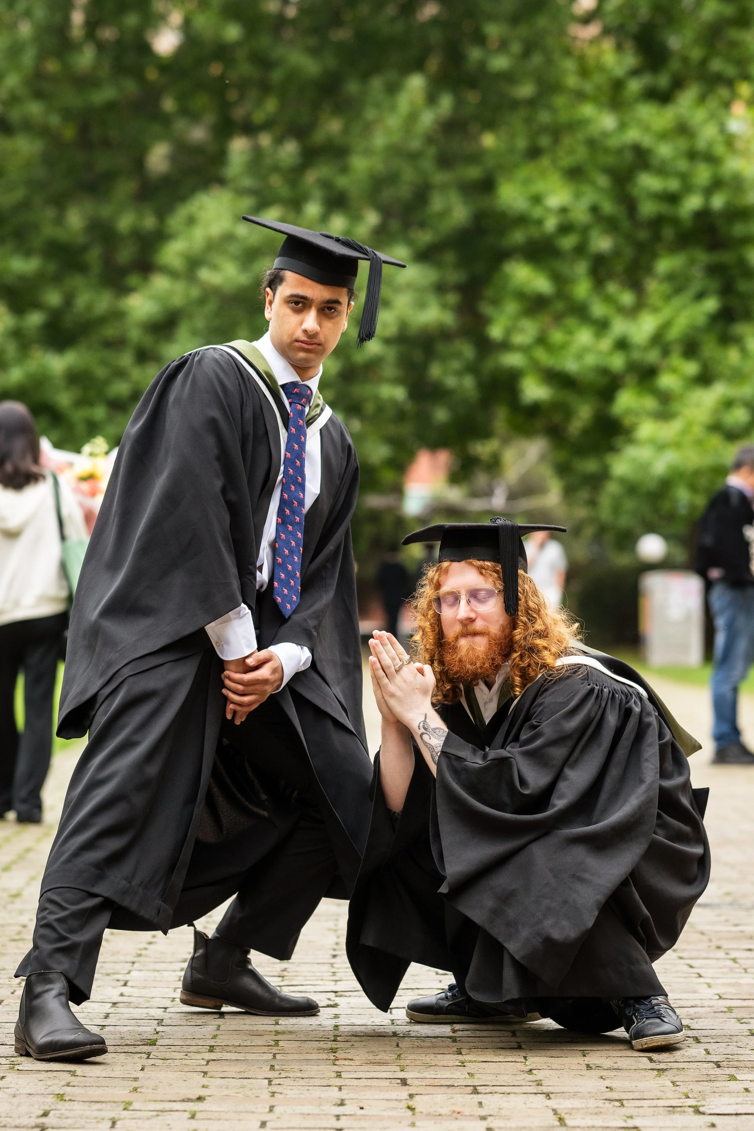 Two graduates in caps and gowns, one standing and one kneeling with hands in a prayer position, outdoors with trees in the background.