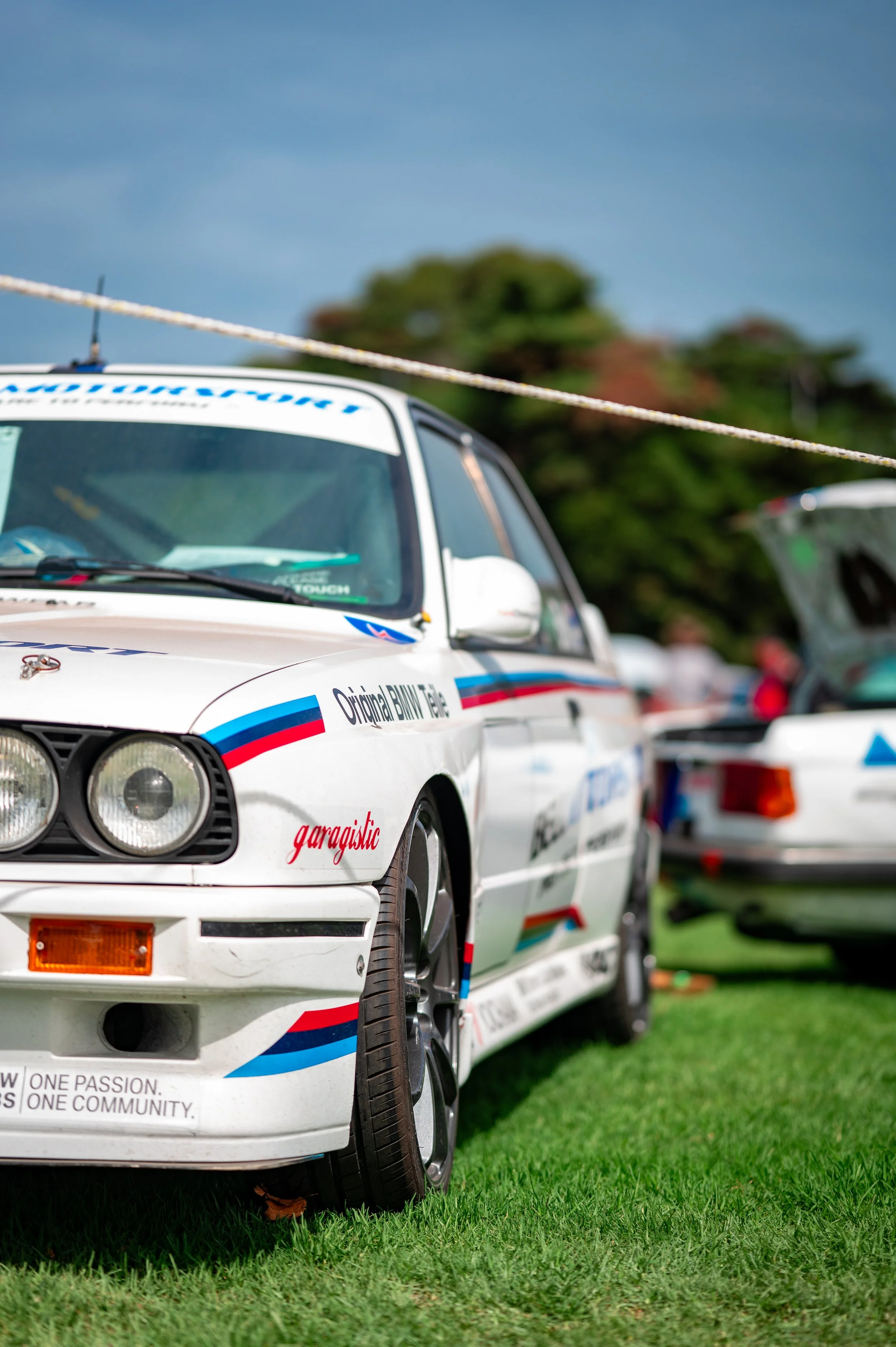 A white BMW race car with blue, red, and black decals parked on grass at an outdoor event.