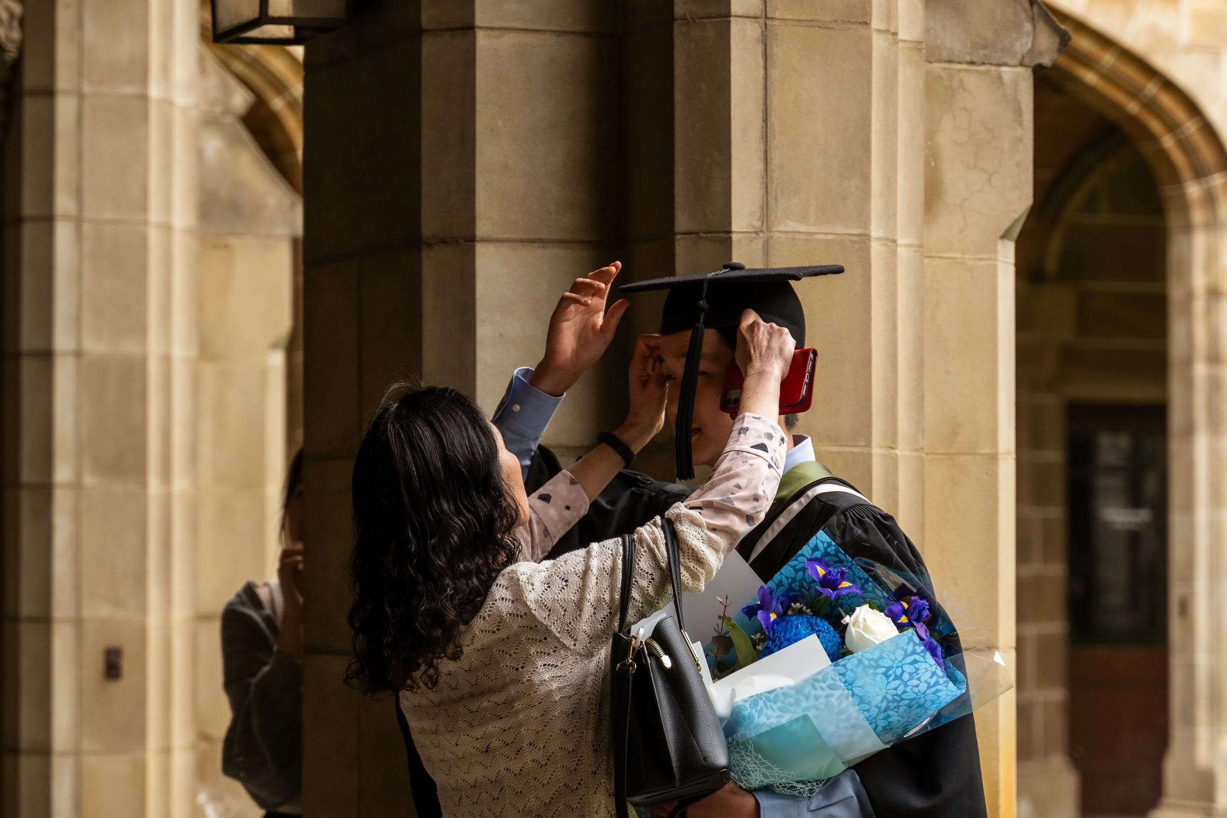 A woman helping a graduate adjust his cap during a graduation ceremony inside a building with stone arches. The graduate is holding a bouquet of flowers.