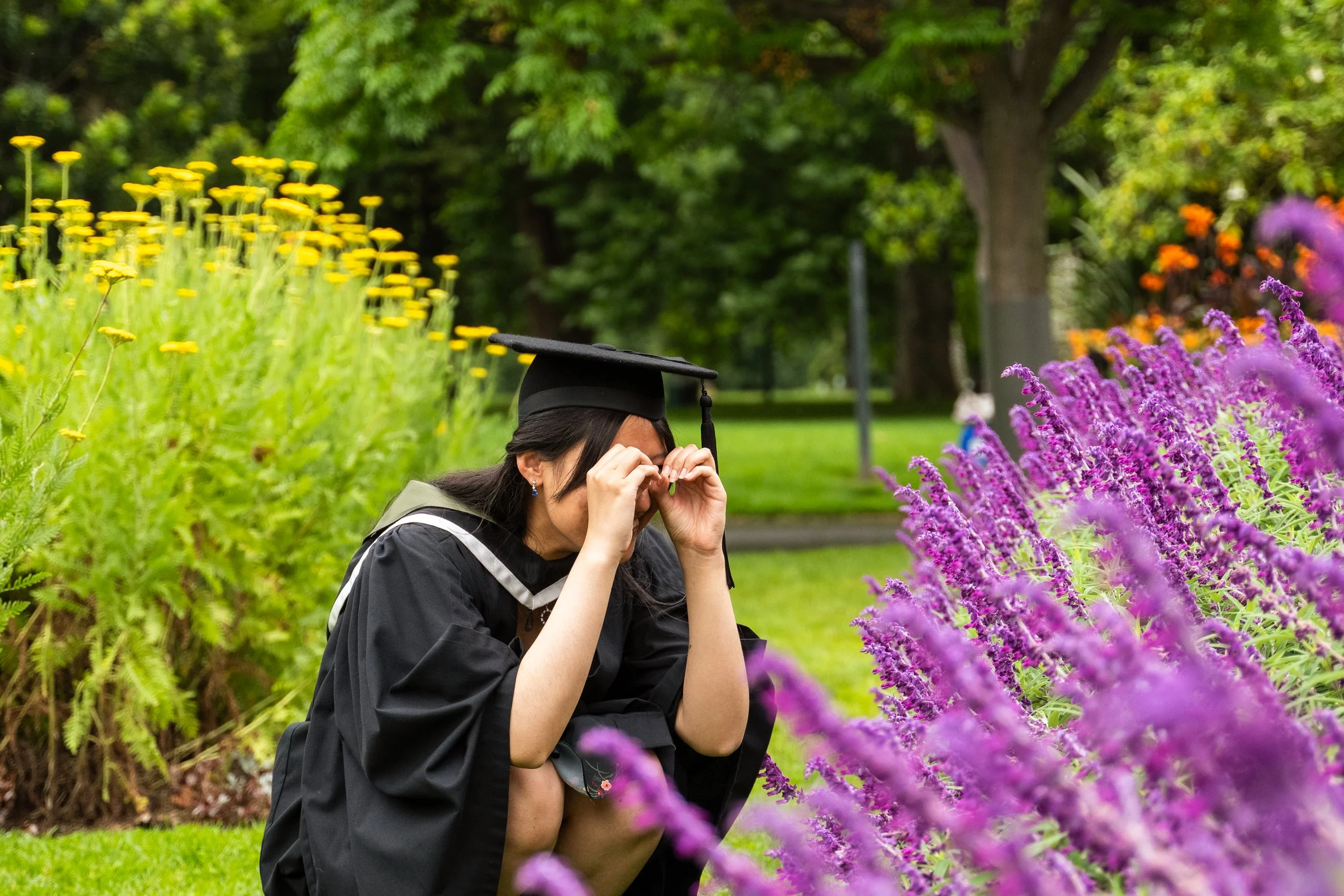 A woman in a graduation cap and gown kneels in a garden, adjusting her glasses, surrounded by purple and yellow flowers and lush green trees.