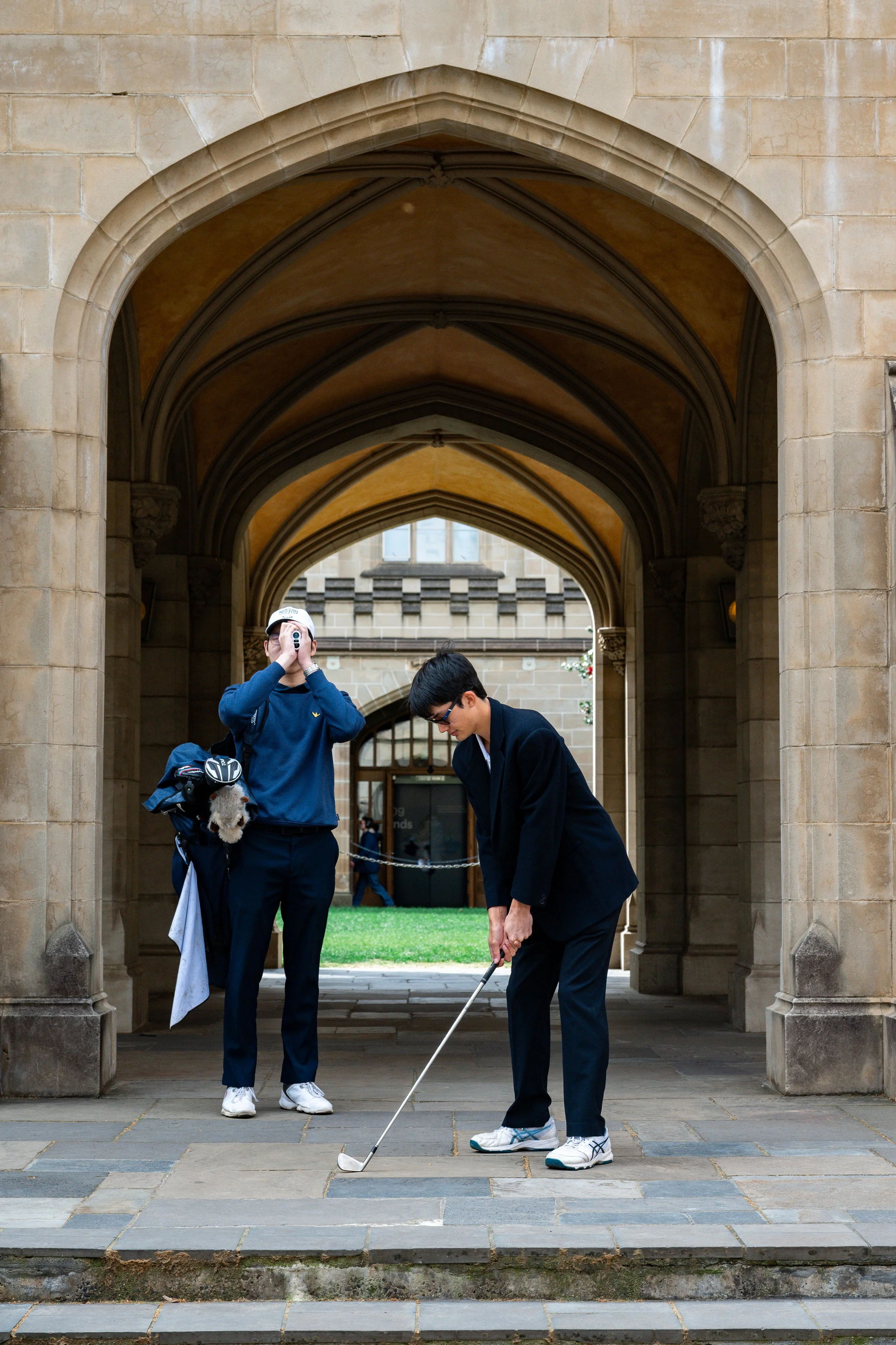 Two young men dressed in sportswear playing mini golf under a stone archway.