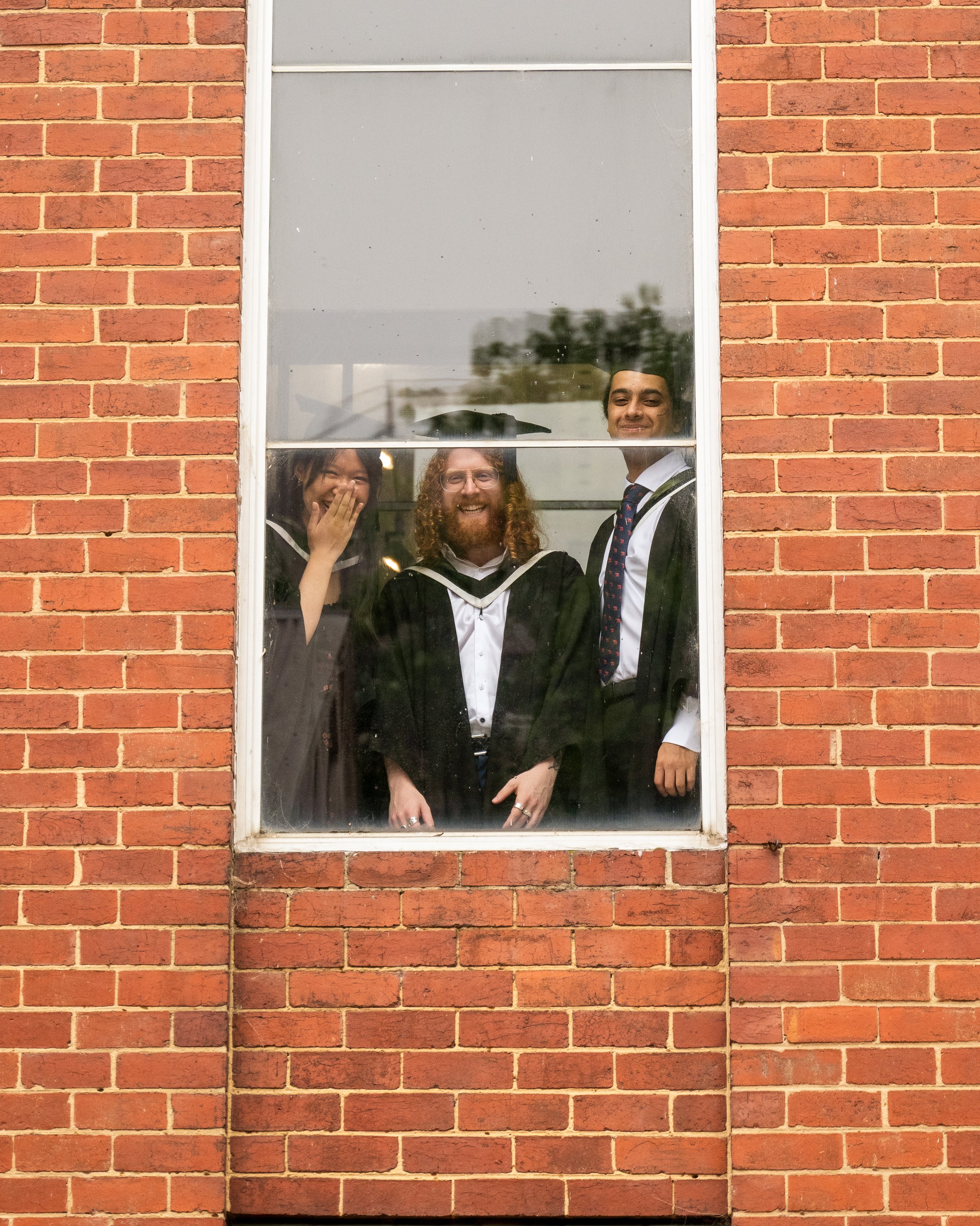 Three graduates in caps and gowns smiling and looking through a window with a brick wall surrounding it.