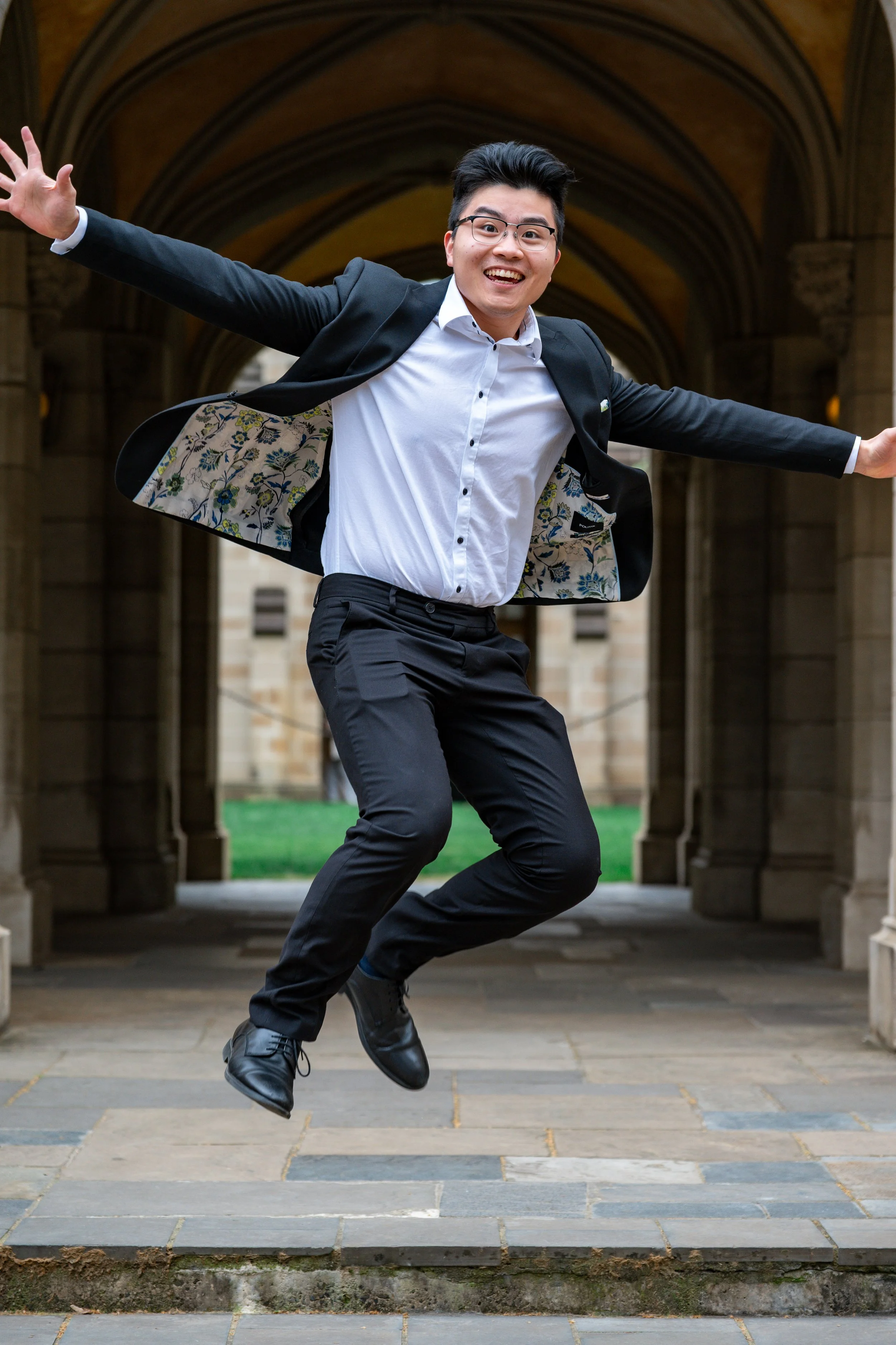 A young man in formal attire jumping with arms wide open in front of a historic stone building.