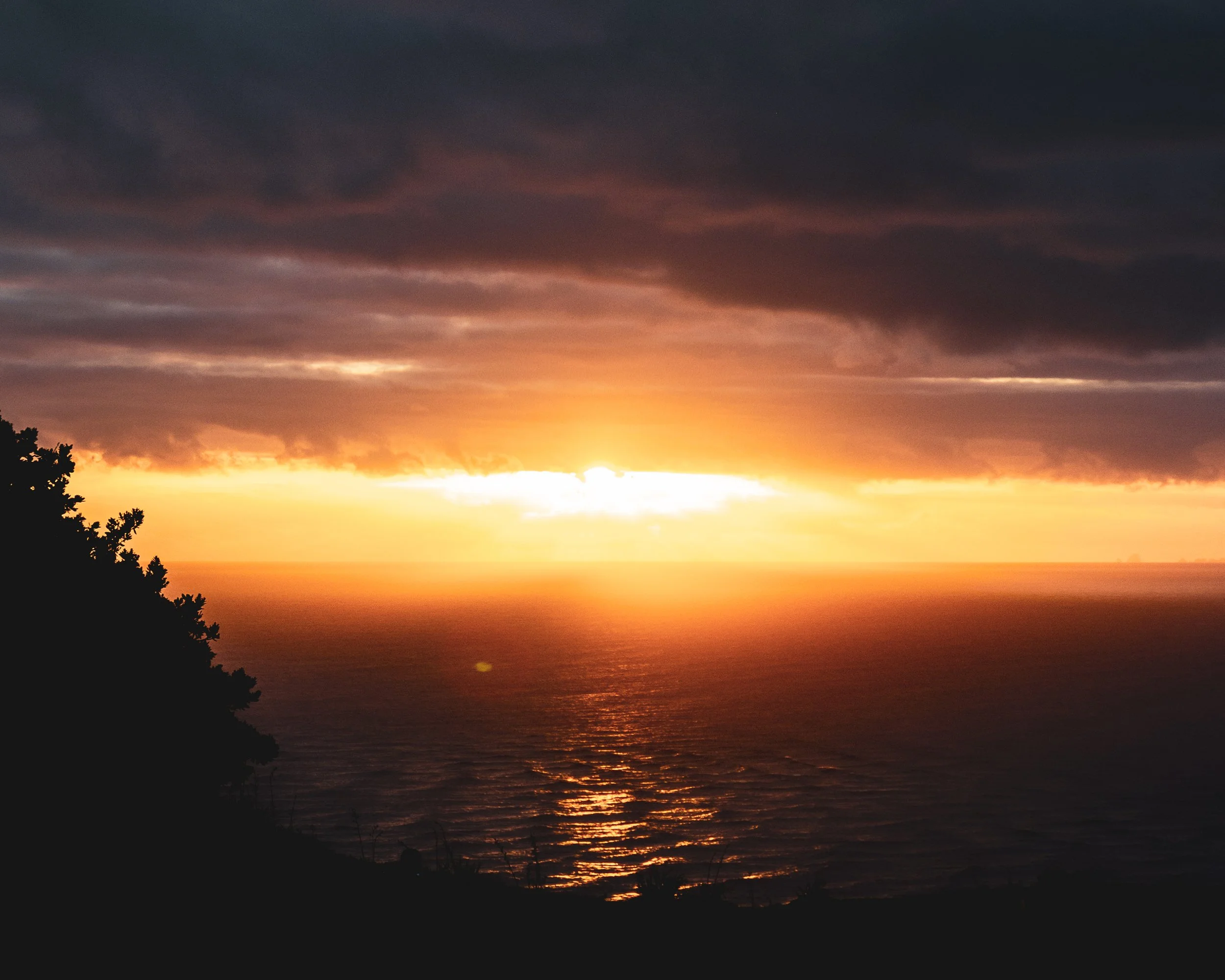 Sunset over the ocean with dark clouds and a silhouette of trees in the foreground.