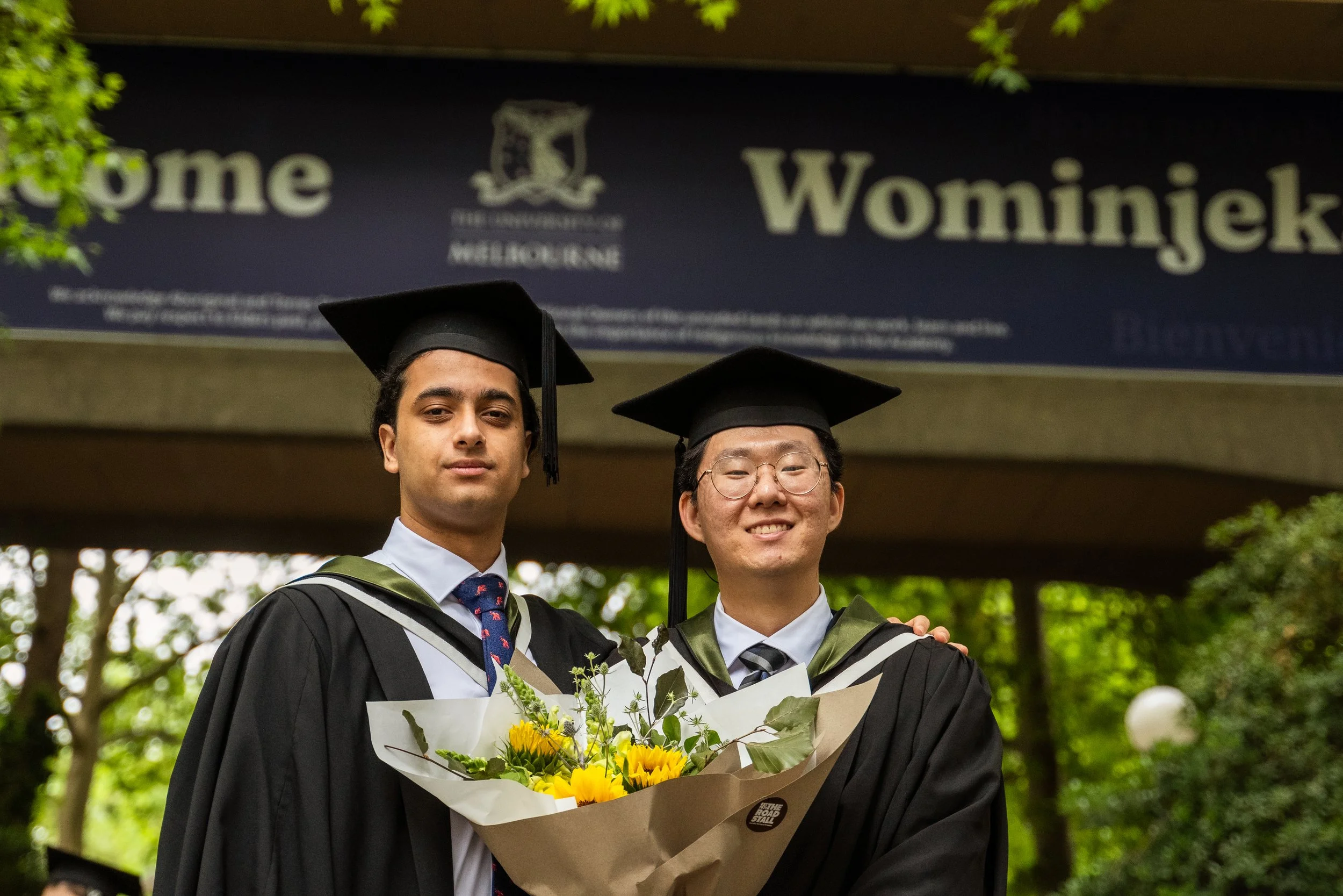Two university graduates in black caps and gowns, one holding a bouquet of yellow flowers, standing outdoors under a sign that reads 'Wominjek' and 'Melbourne' in the background.