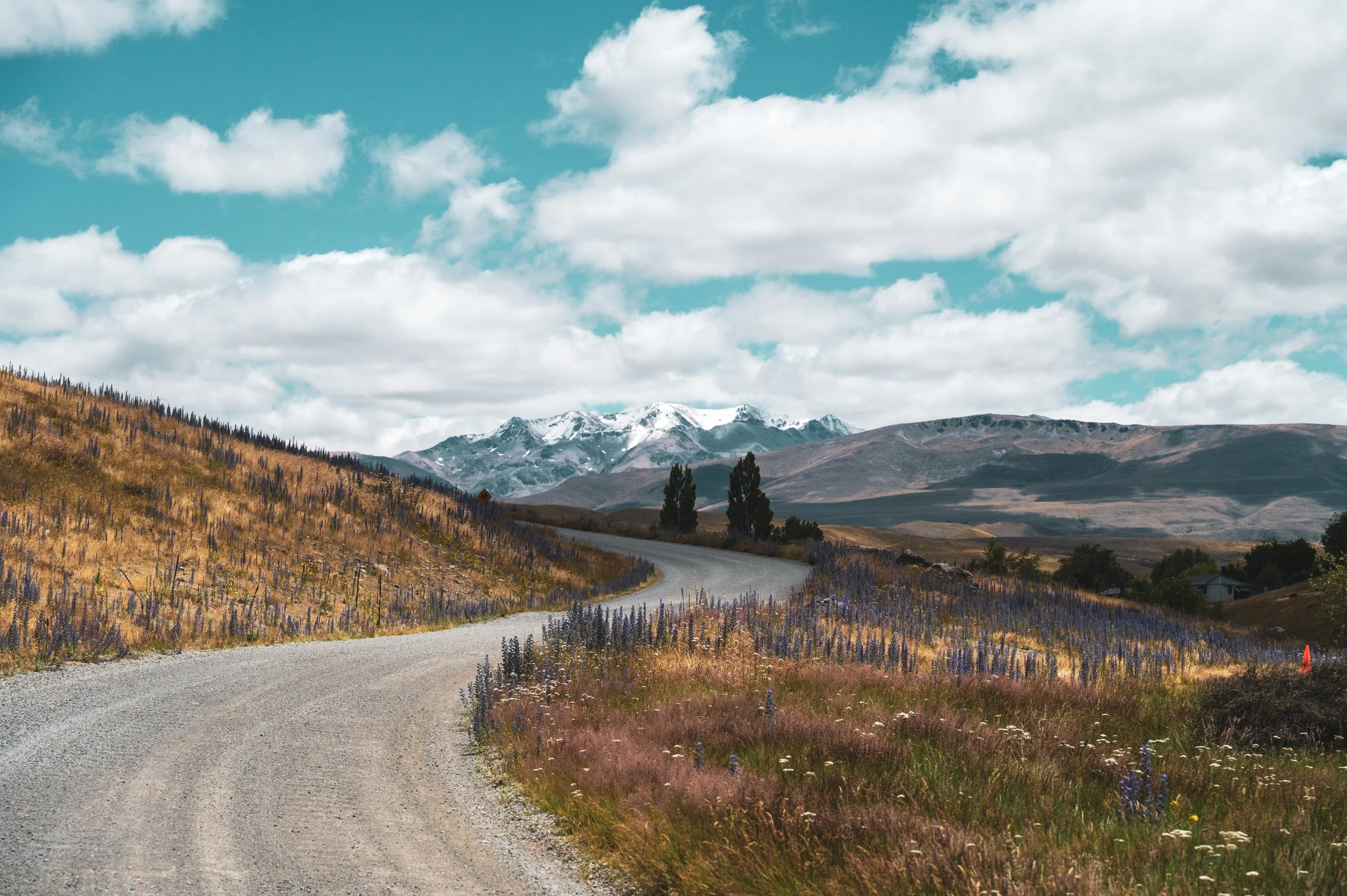A winding dirt road in a rural landscape with mountains in the background and a partly cloudy sky.