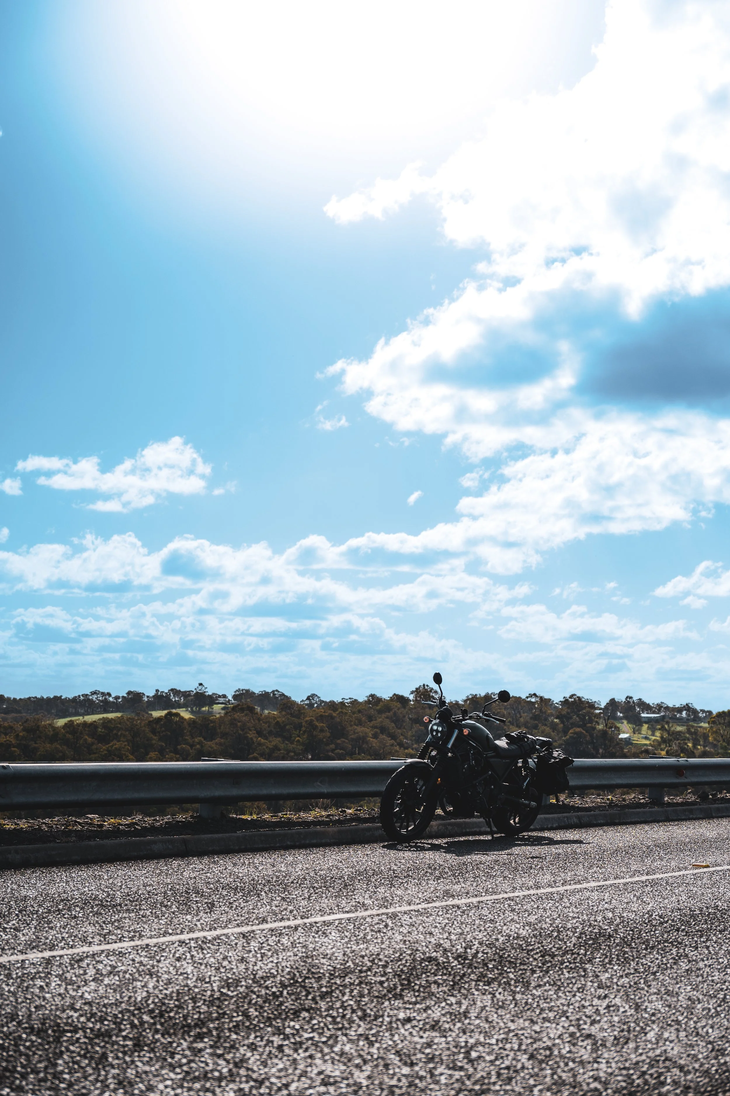 Black motorcycle parked on the side of a rural road with guardrail, open landscape, trees, blue sky, and clouds overhead.