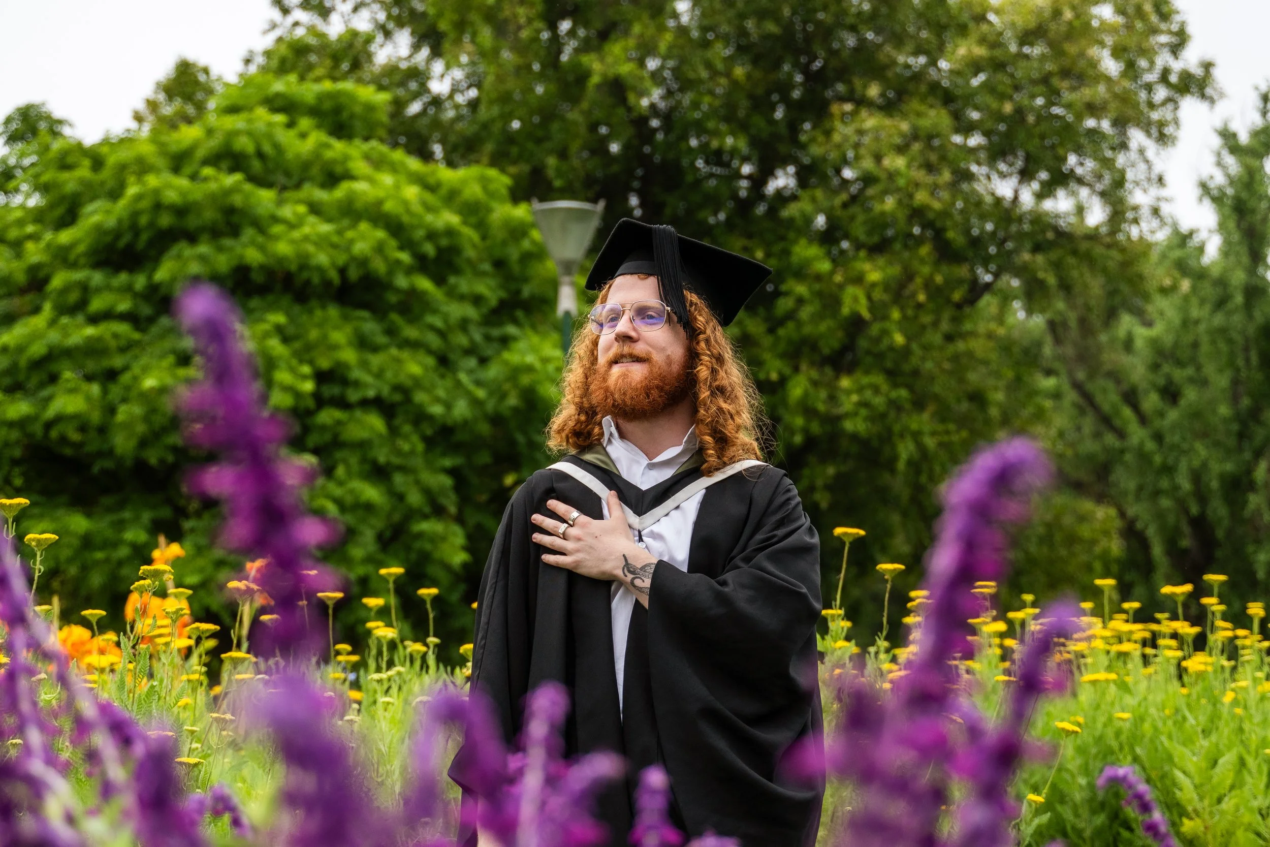 A man in graduation attire, wearing a cap and gown, standing outside in a field of purple and yellow flowers with Green trees in the background.
