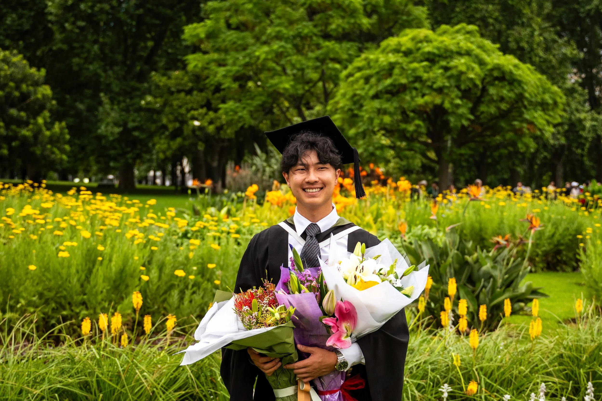 Young man in graduation cap and gown smiling while holding a bouquet of flowers in a garden with yellow and orange flowers and green trees.