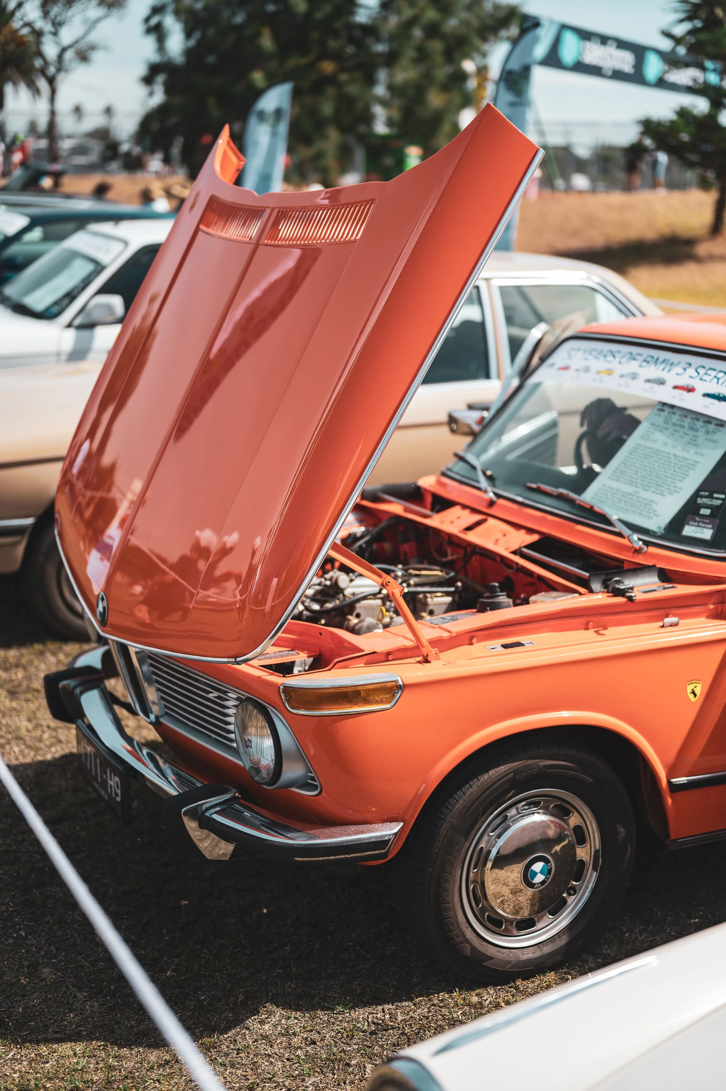Orange vintage BMW car with the hood open, parked at a car show with other cars and spectators in the background.