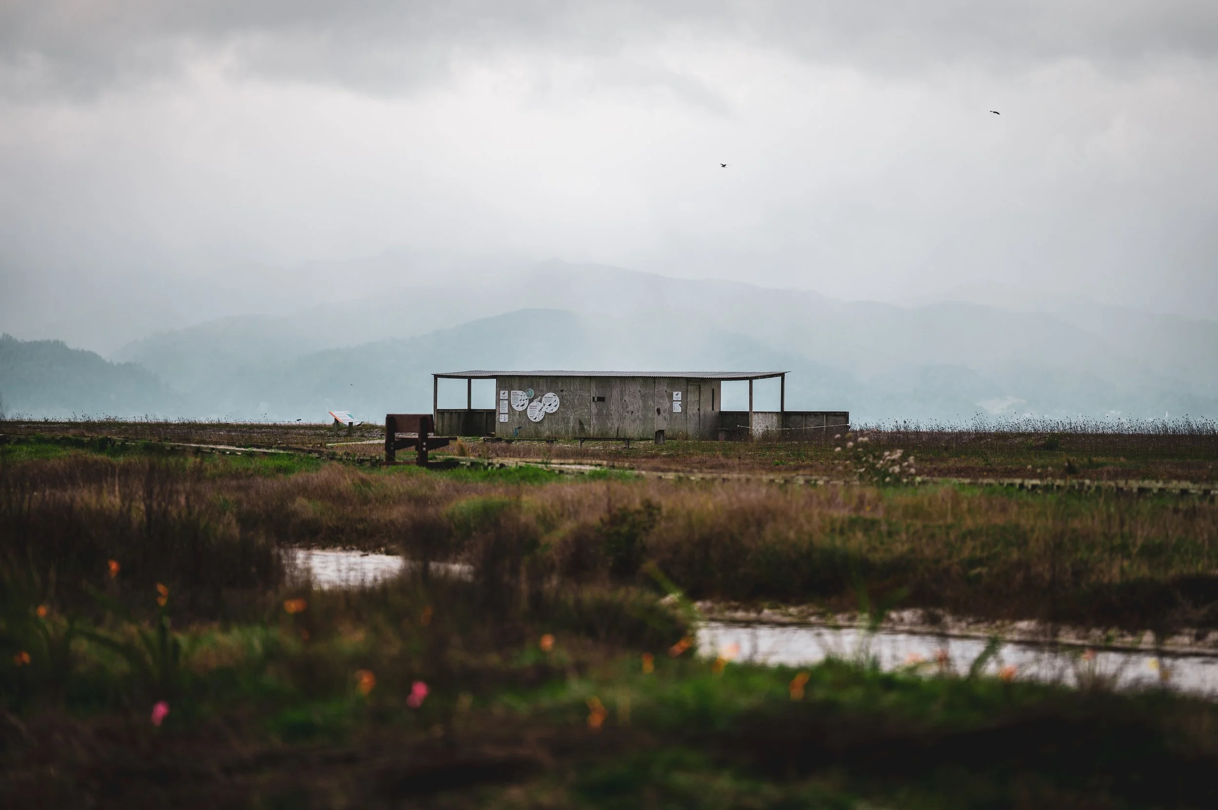 An abandoned, weathered wooden structure on a grassy field with a bench nearby, under a cloudy sky with mountains in the background.